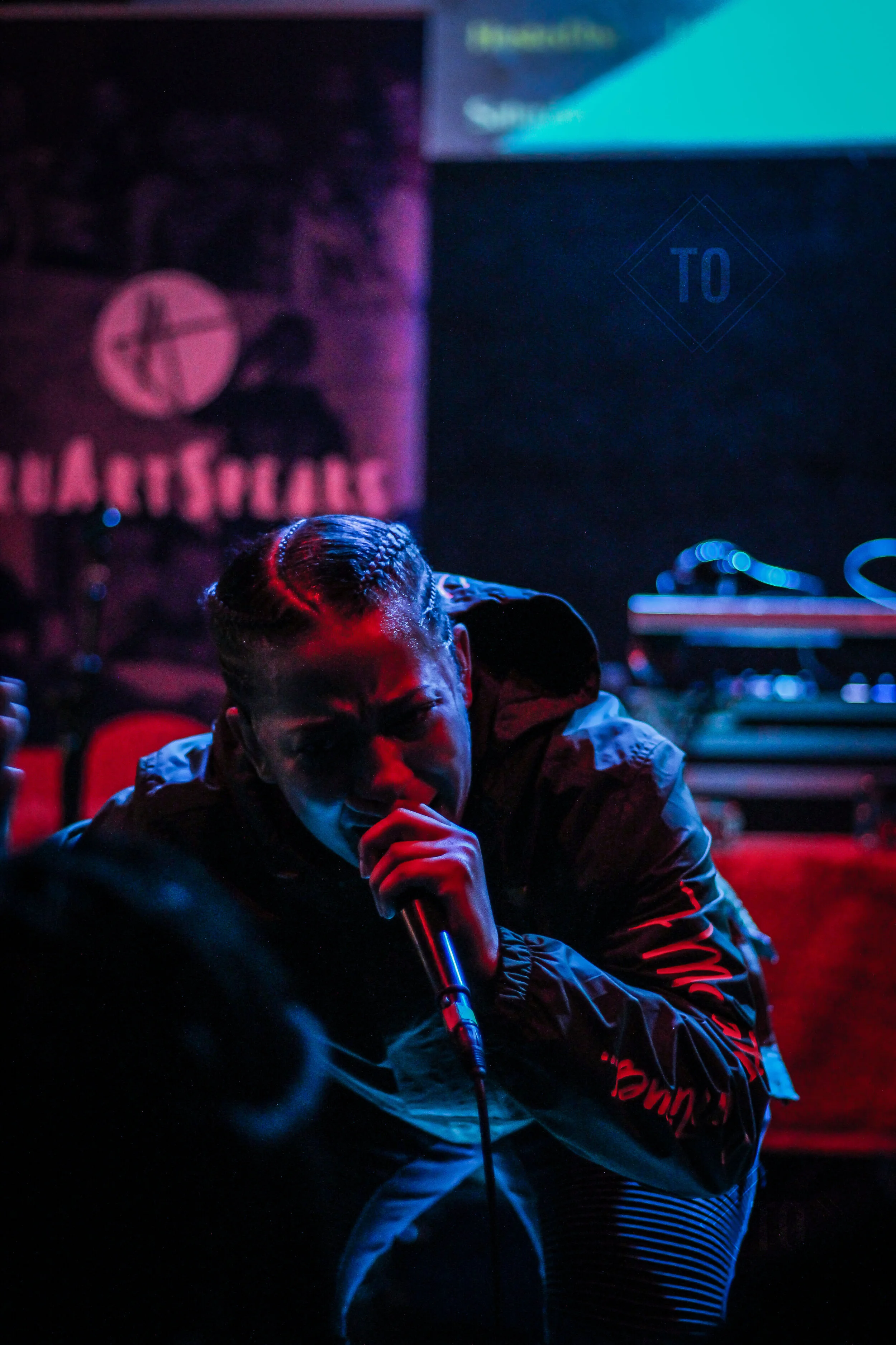 A person with braided hair singing passionately into a microphone on a dimly lit stage with colorful red and blue lighting, and DJ equipment visible in the background.