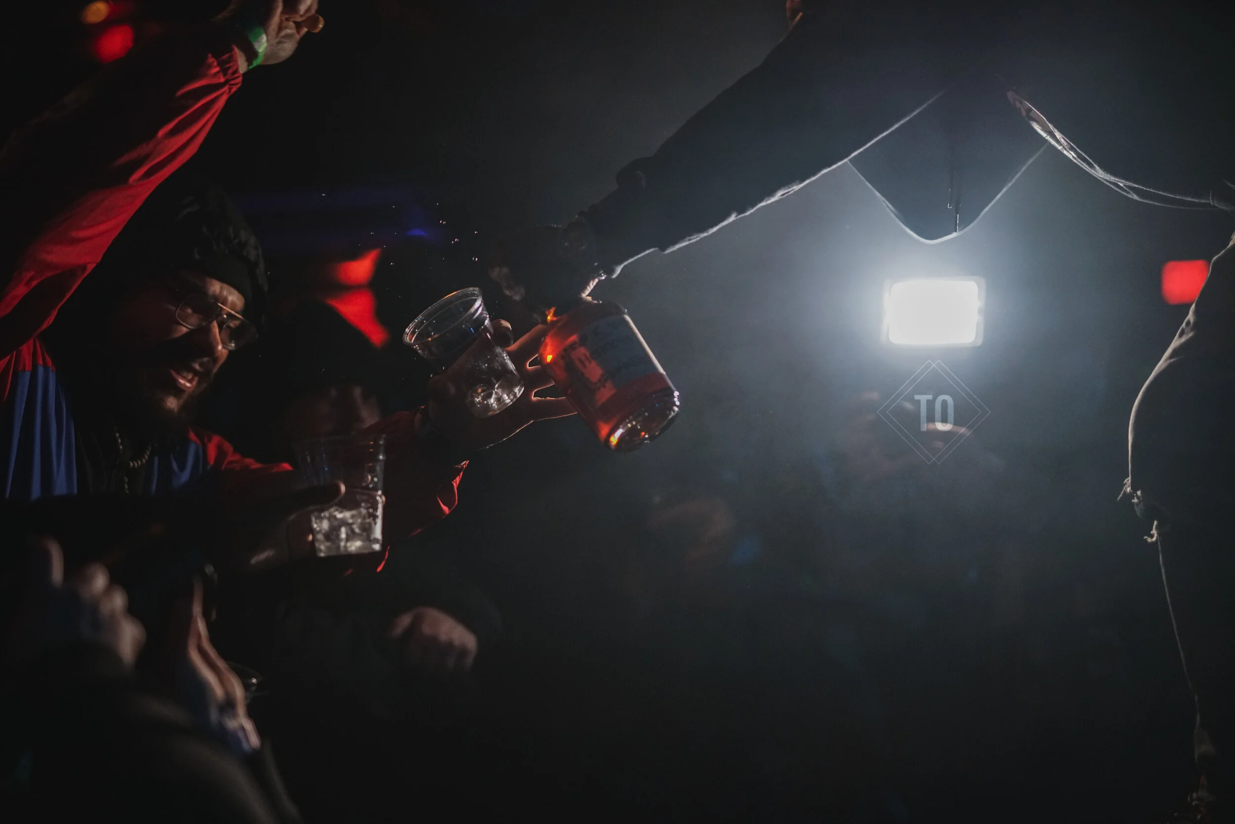 People raising glasses of alcohol in a dark, lively setting, with a bright light in the background.