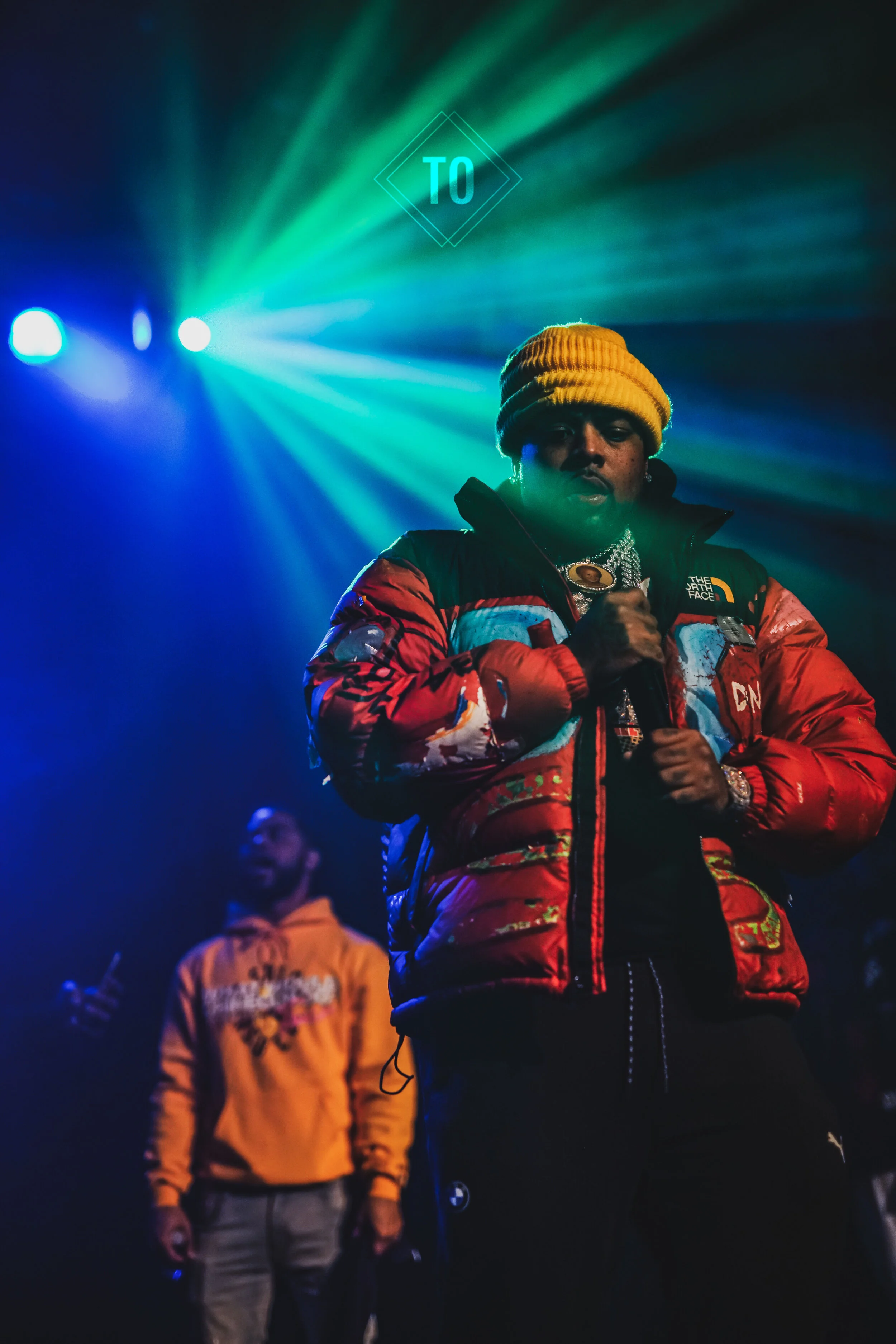 A person in a red winter jacket and yellow beanie holding a microphone, illuminated by colorful stage lights, with a blurred person in a yellow hoodie in the background.