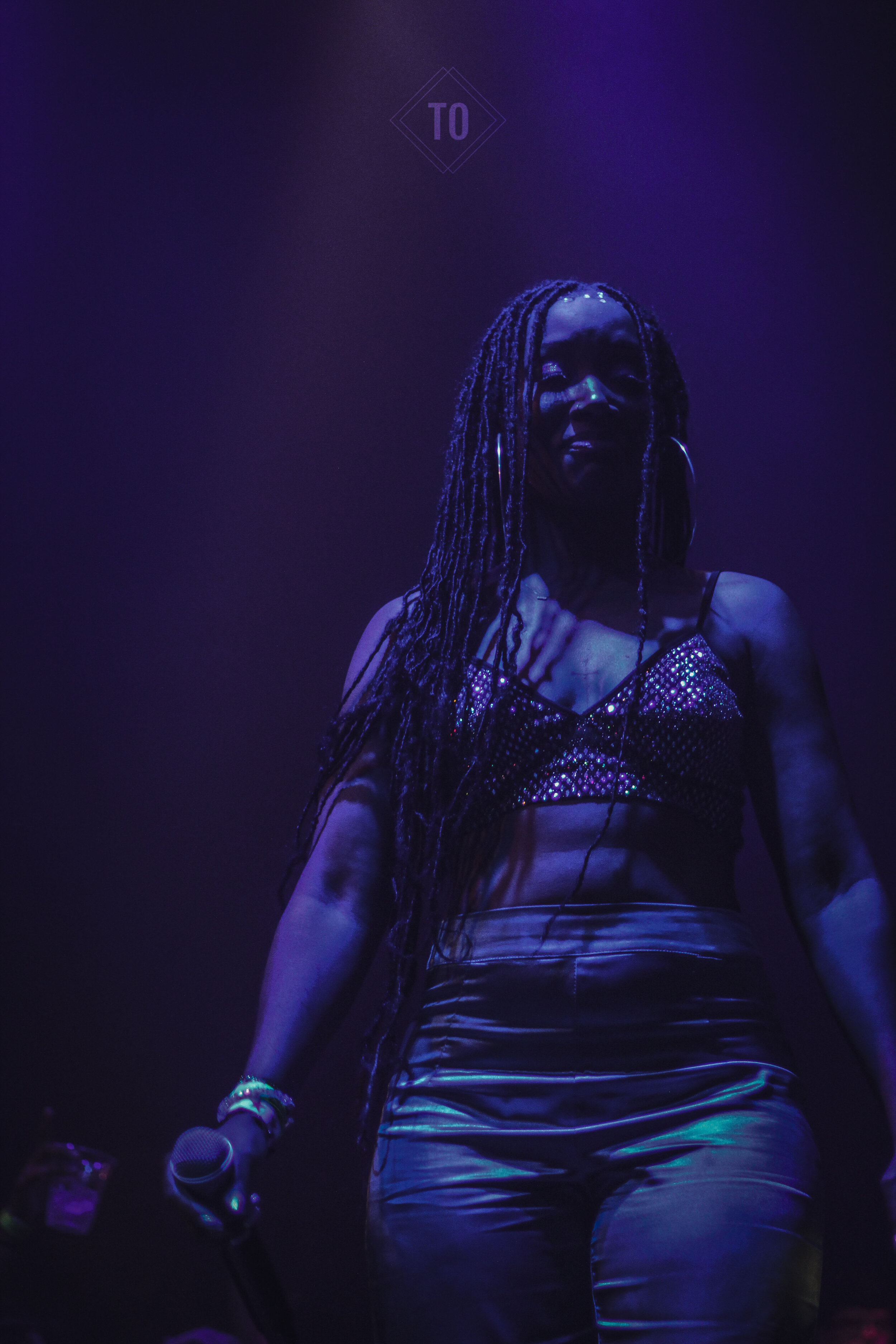A woman with long dreadlocks holding a microphone, illuminated by purple stage lights, wearing a sparkly top and tight pants.