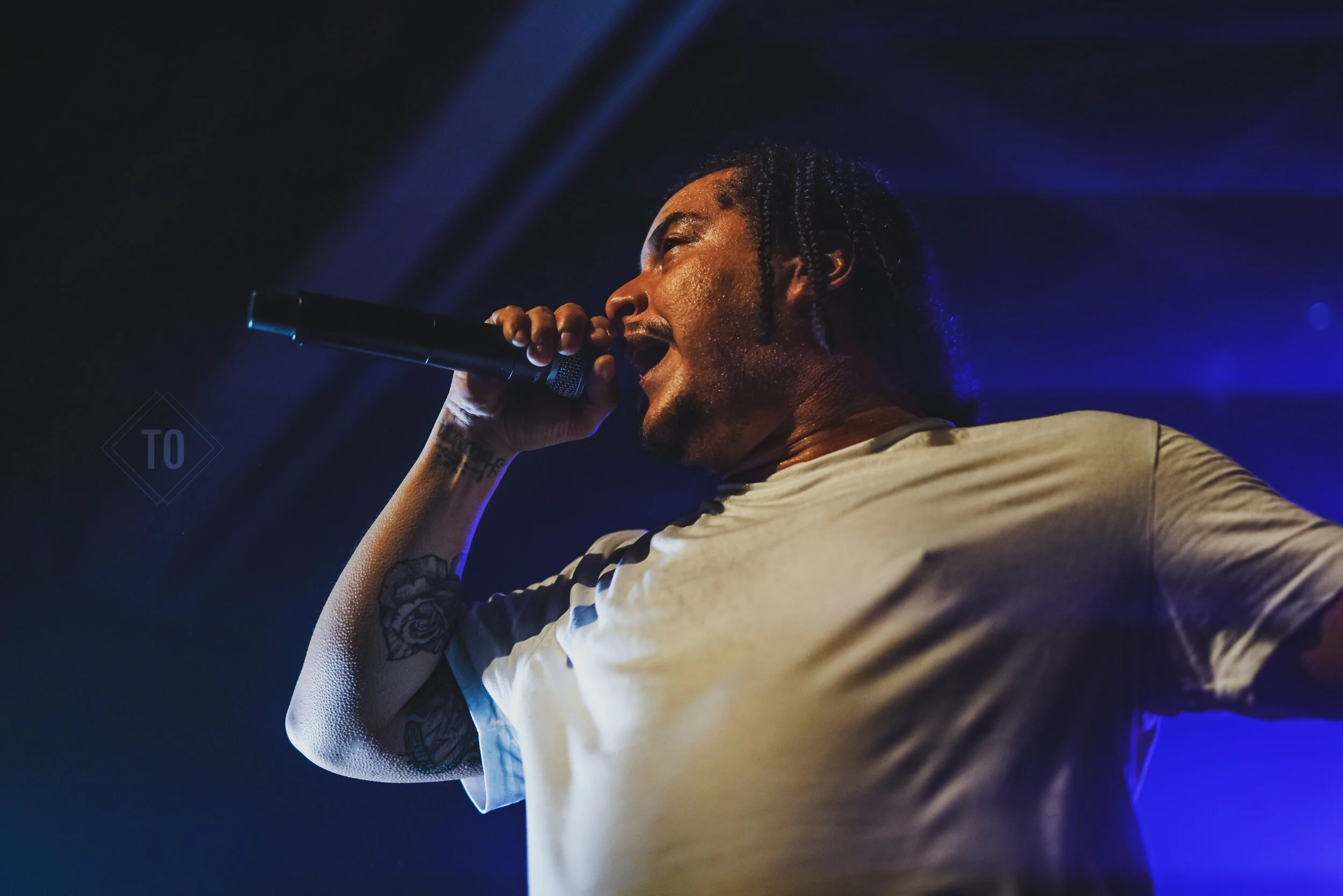 Male performer singing into a microphone on stage, with visible tattoos on his arm, dark background, blue stage lighting.