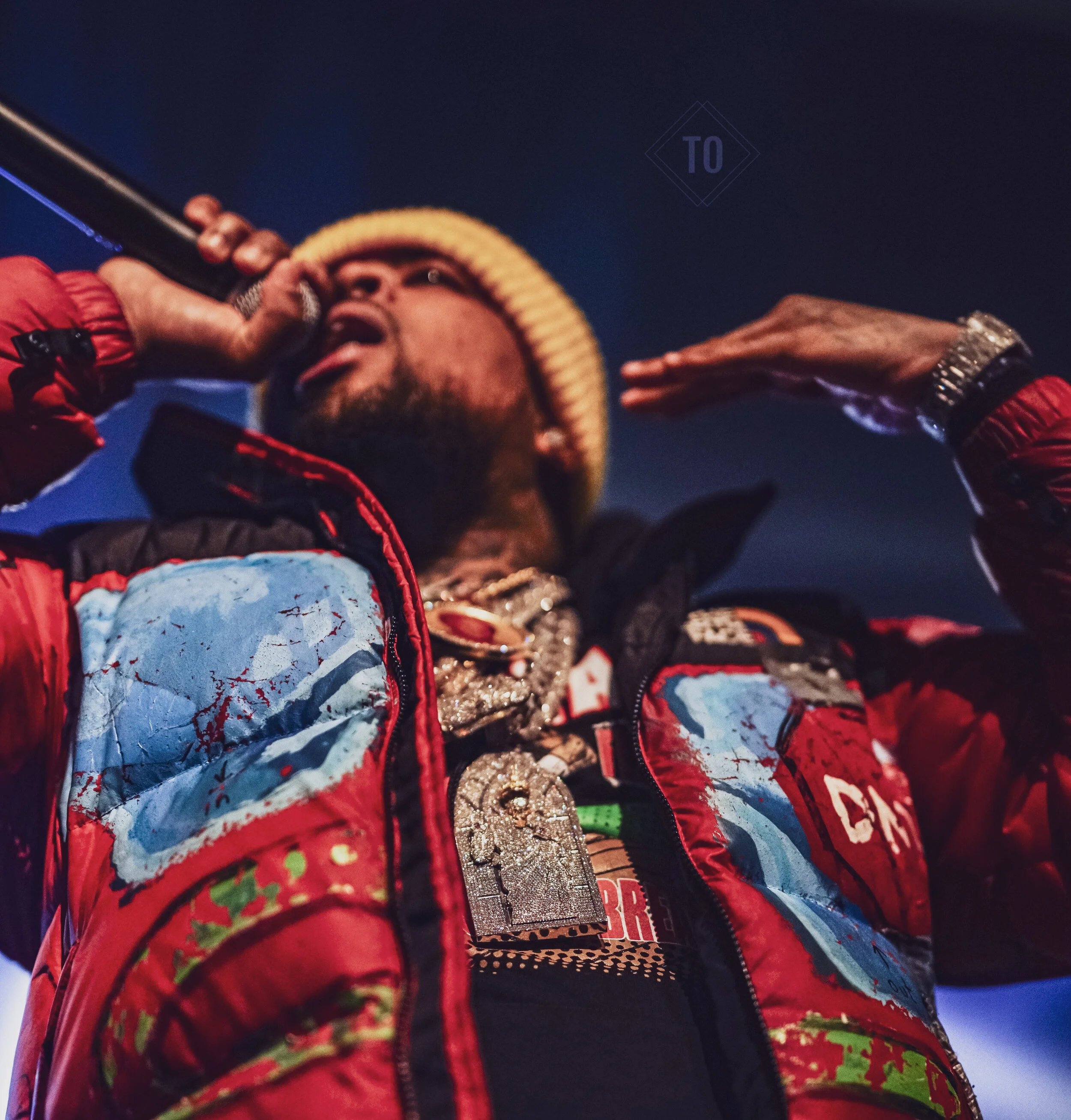 A person wearing a beanie and a red jacket with patches and chains is singing or speaking into a microphone. The person is making a saluting gesture with one hand near the head, with a dark sky in the background.