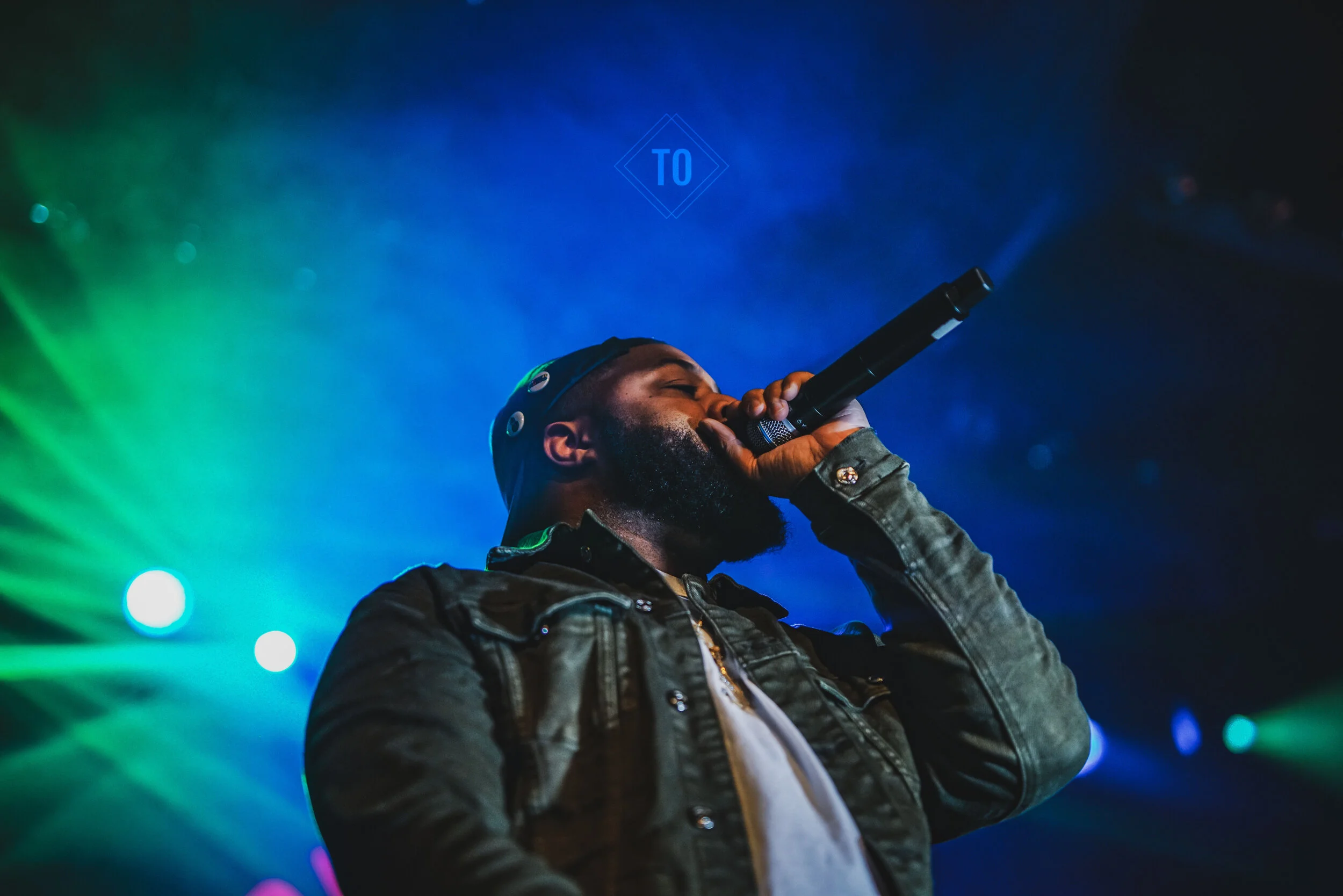 A man with a beard wearing a dark jacket and a cap singing into a microphone on stage with colorful stage lighting.