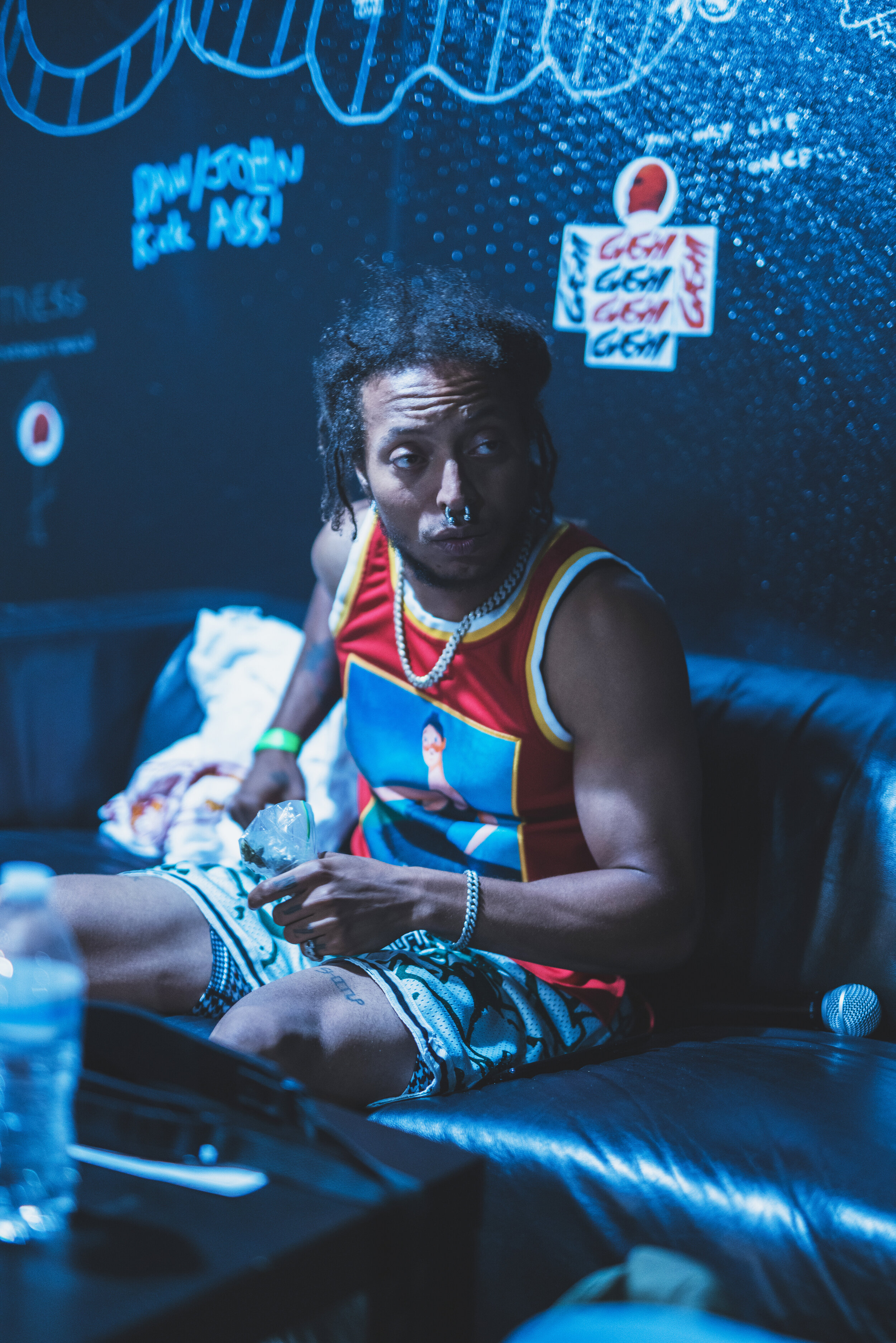A young man with dreadlocks and piercings, wearing a colorful graphic tank top and patterned shorts, sitting on a black leather couch in a dimly lit room decorated with posters and graffiti, holding a plastic bag and a water bottle.