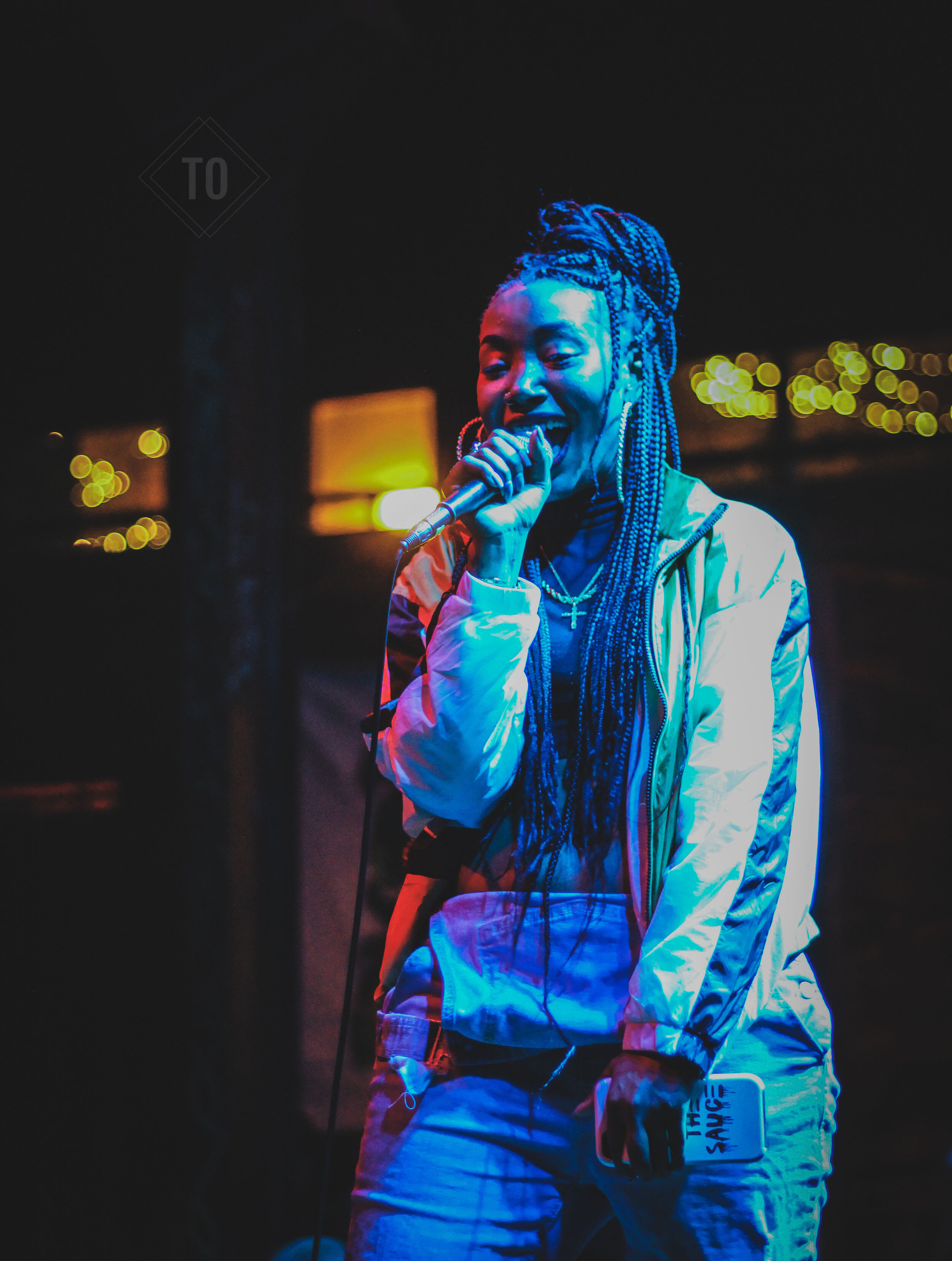 A woman singing into a microphone at night, with colorful lighting and blurred city lights in the background.