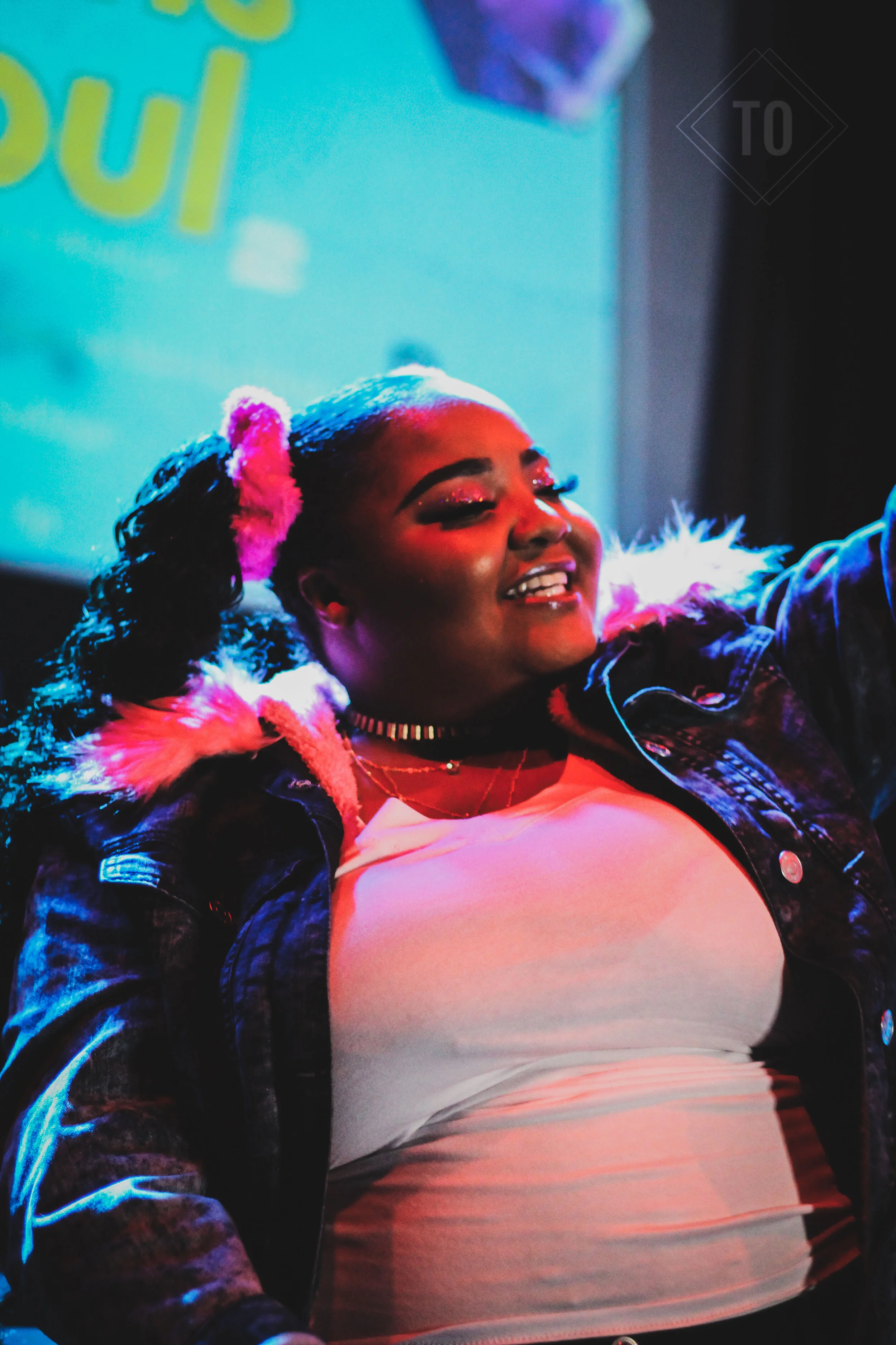 A plus-sized woman with long braided hair in a colorful ponytail, smiling and posing with one arm raised. She is wearing a white top, a choker necklace, and a black leather jacket with faux fur-lined hood. The background features a digital screen wit