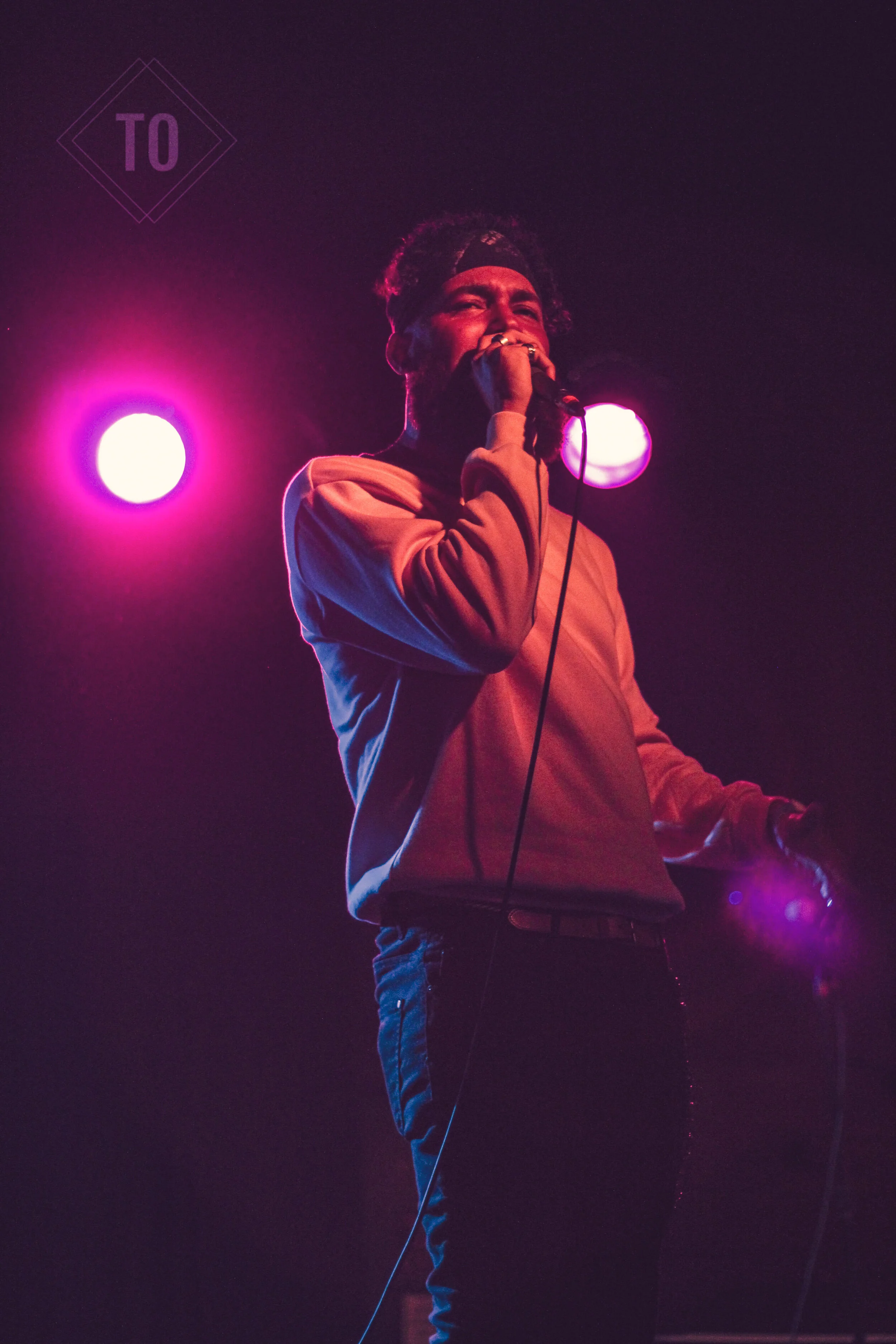 A male performer singing or speaking into a microphone on stage, illuminated by colorful pink and purple stage lights.