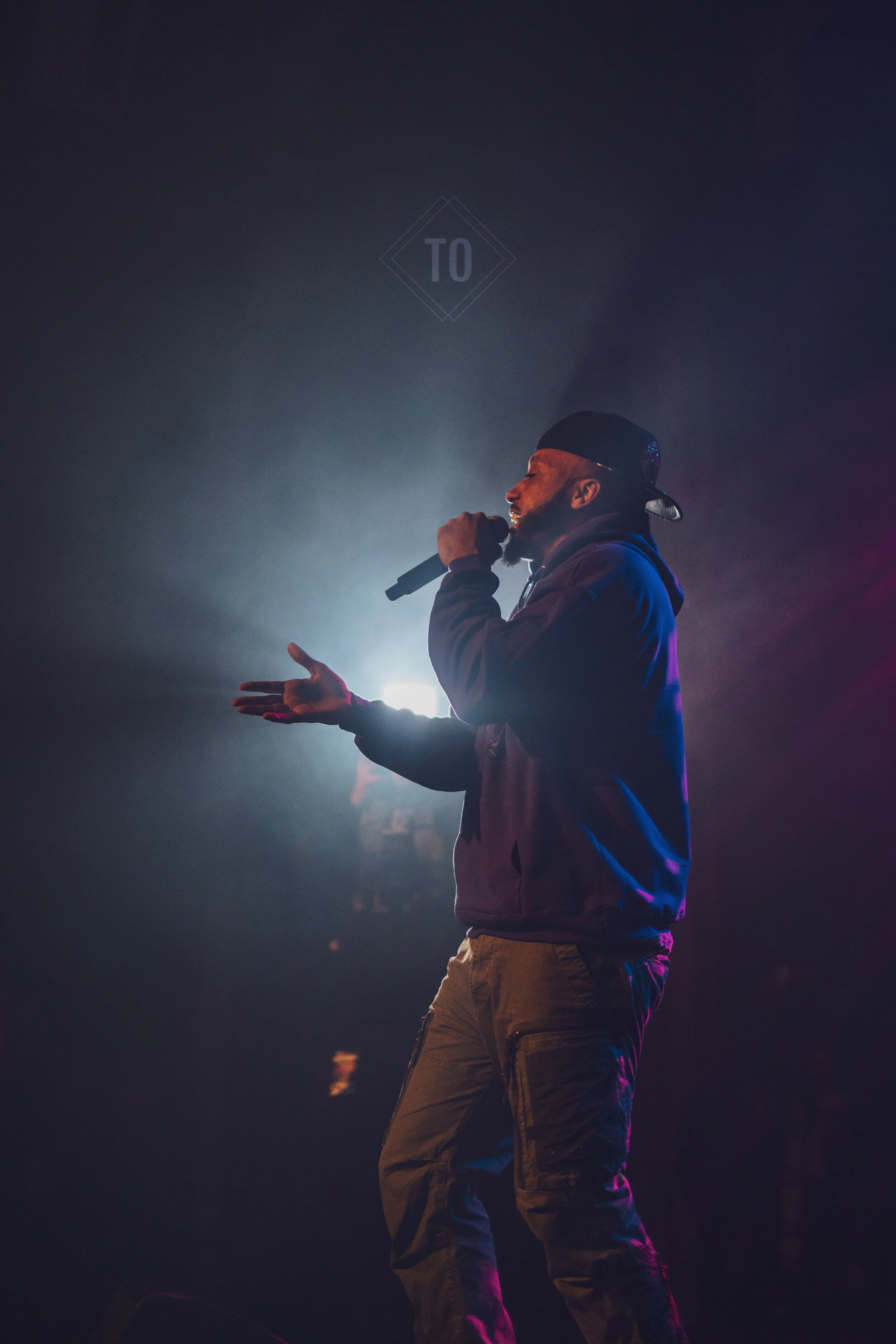 A male rapper wearing a black cap and jacket performs on stage with a microphone, backlit by bright stage lights.