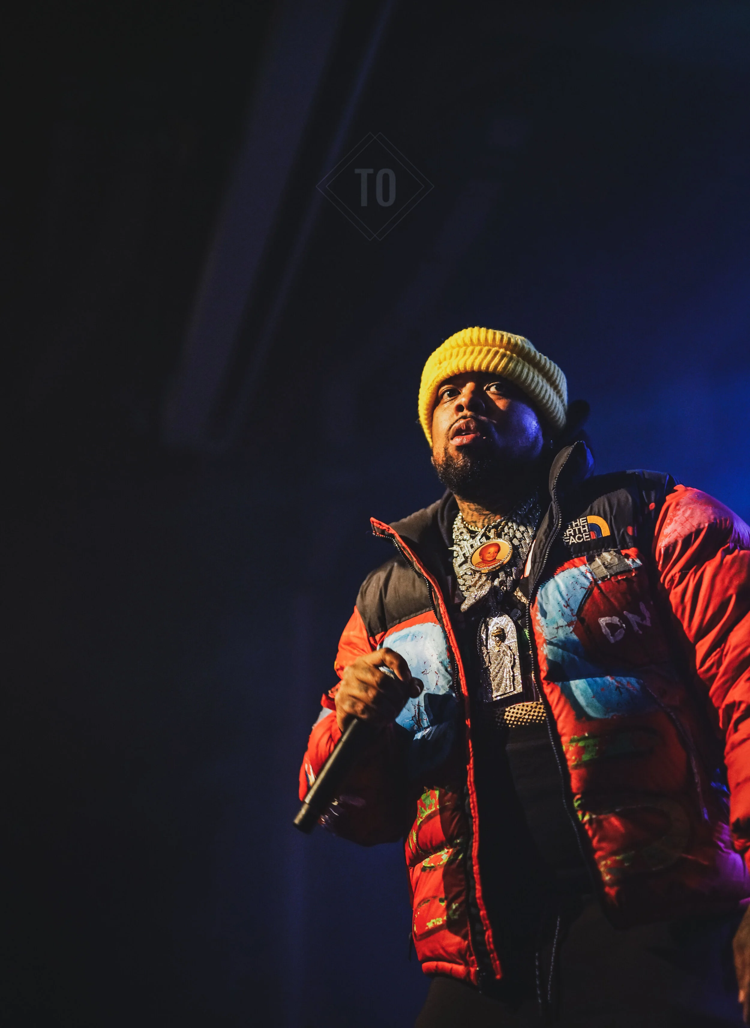 A male performer on stage wearing a yellow knit beanie, a colorful puffer jacket, and multiple necklaces, holding a microphone and looking upwards.