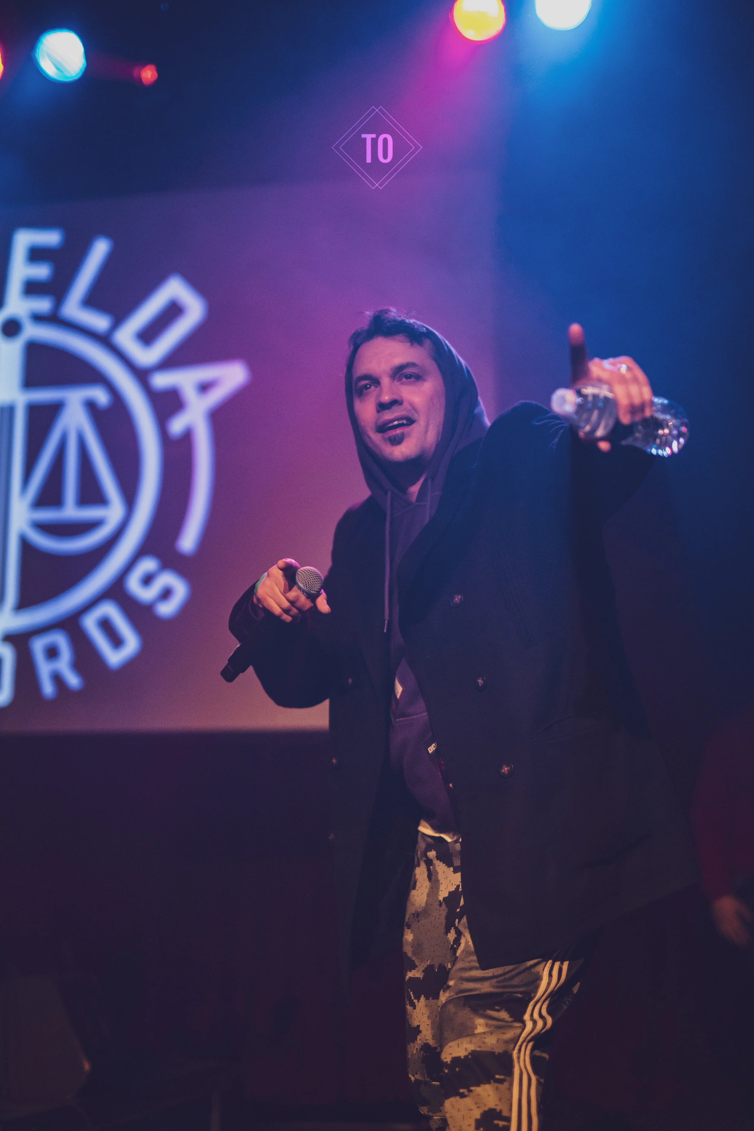 A man on stage holding a microphone in one hand and a water bottle in the other, giving a thumbs up, with a logo that reads 'Lola Awards' in the background, colorful stage lights overhead.