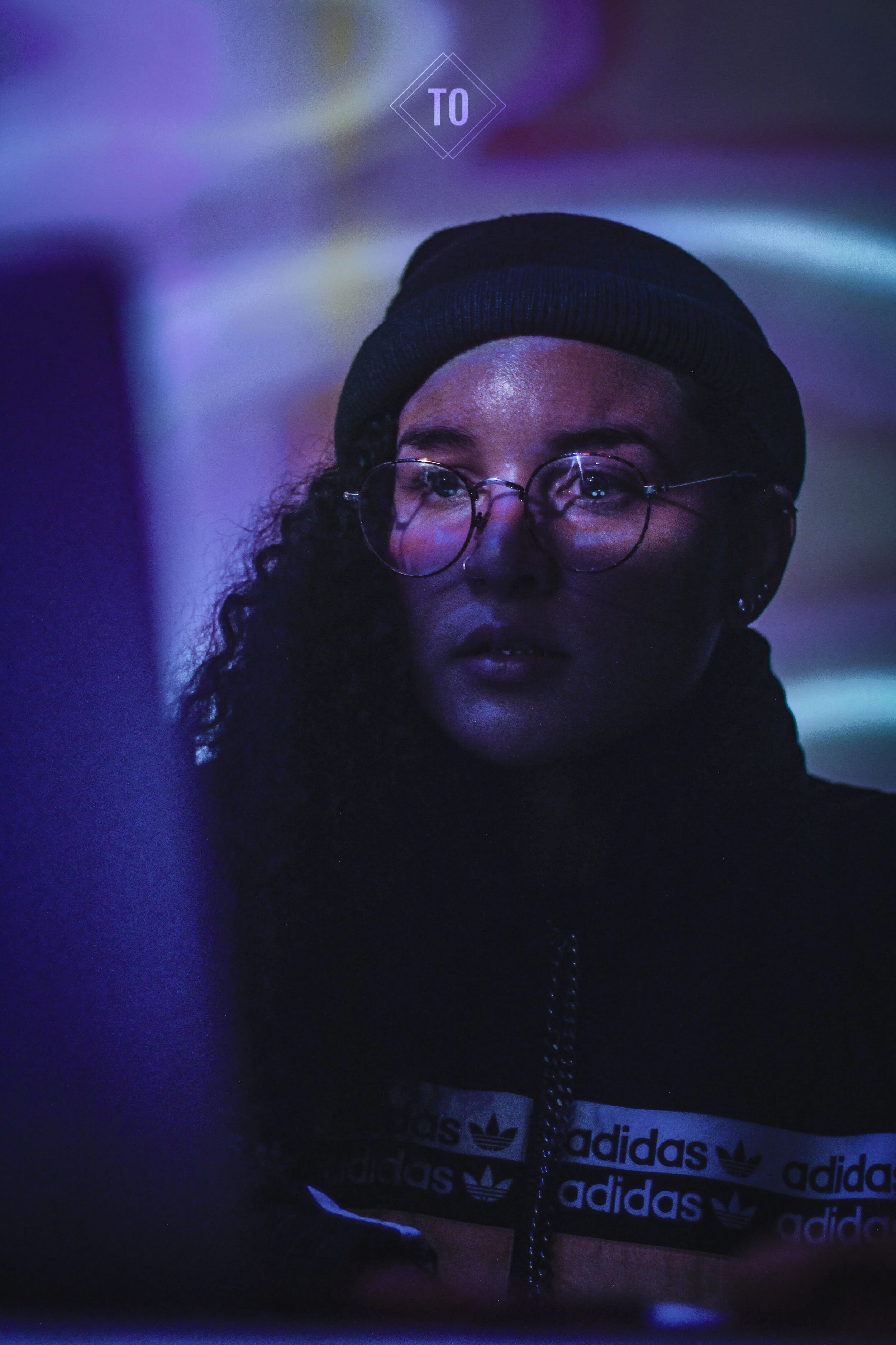 A young woman with curly hair, glasses, and wearing a black beanie looks at something illuminated by blue and purple neon lights.
