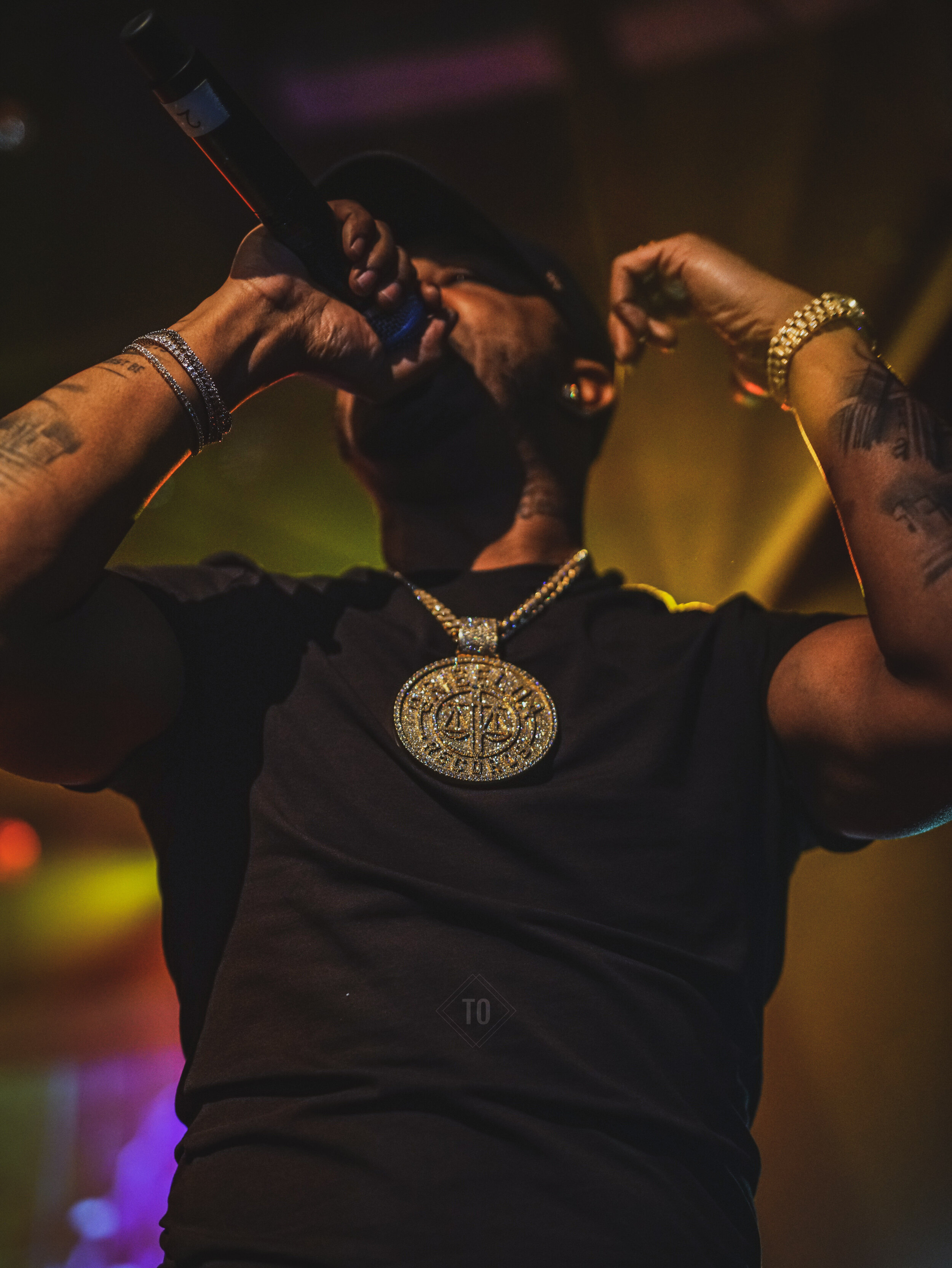 A rapper performing on stage, holding a microphone to his mouth with his right hand, wearing a black cap, black shirt, gold chain with a large medallion, and gold jewelry on his left wrist. The background has colorful stage lights.