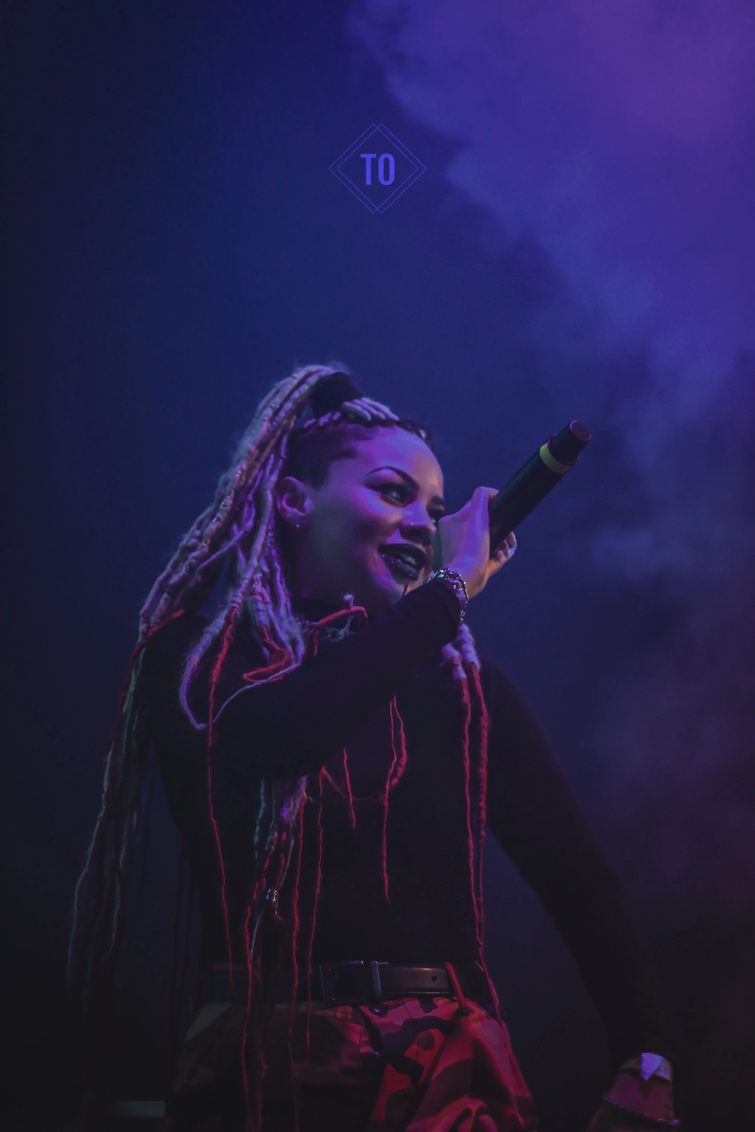A female performer with long dreadlocks singing into a microphone on stage with purple and blue lighting.
