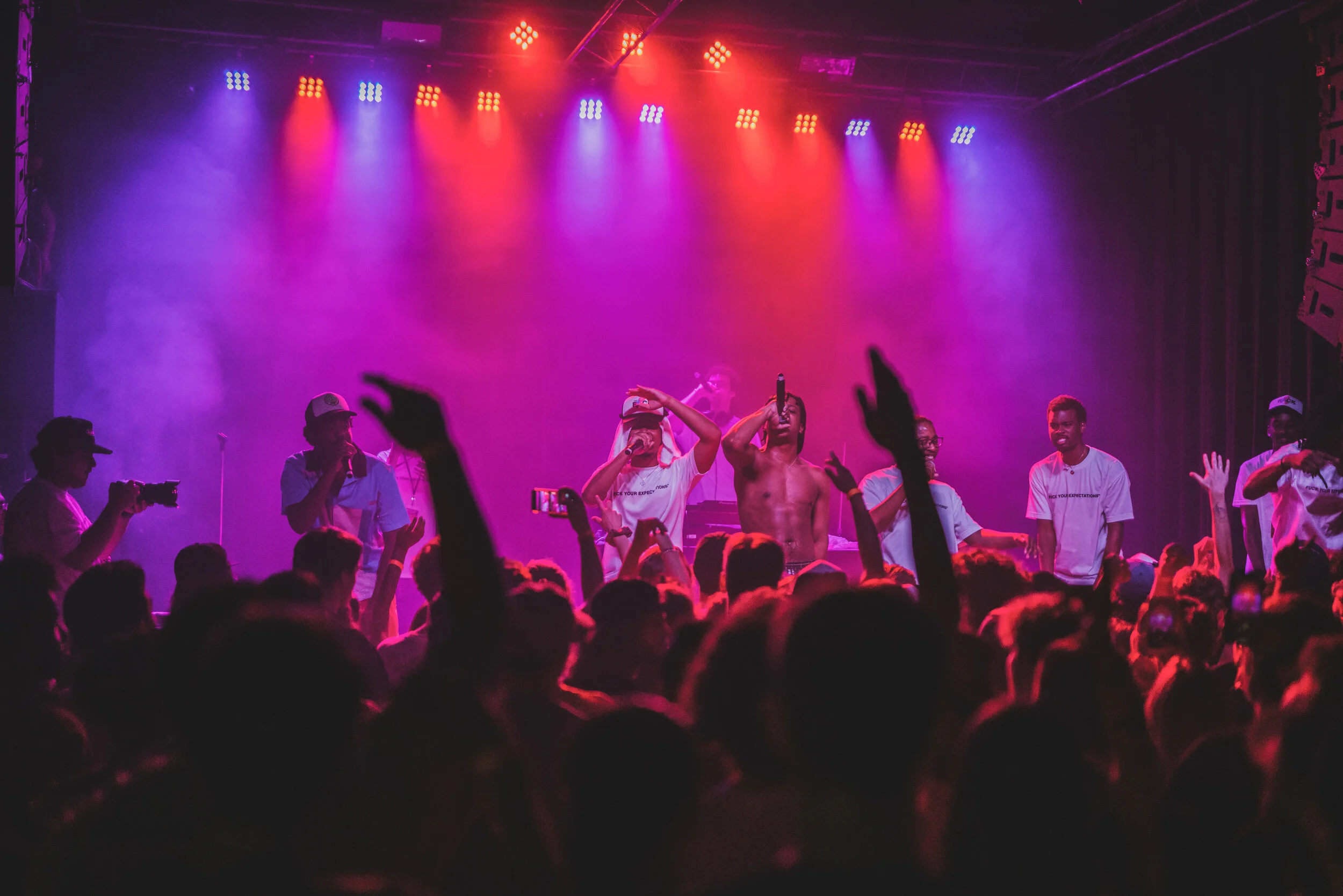 A concert scene with a crowd dancing and raising their hands. Performers on stage include a shirtless man holding a microphone, others wearing white shirts, with colorful stage lighting including purple, red, and blue.