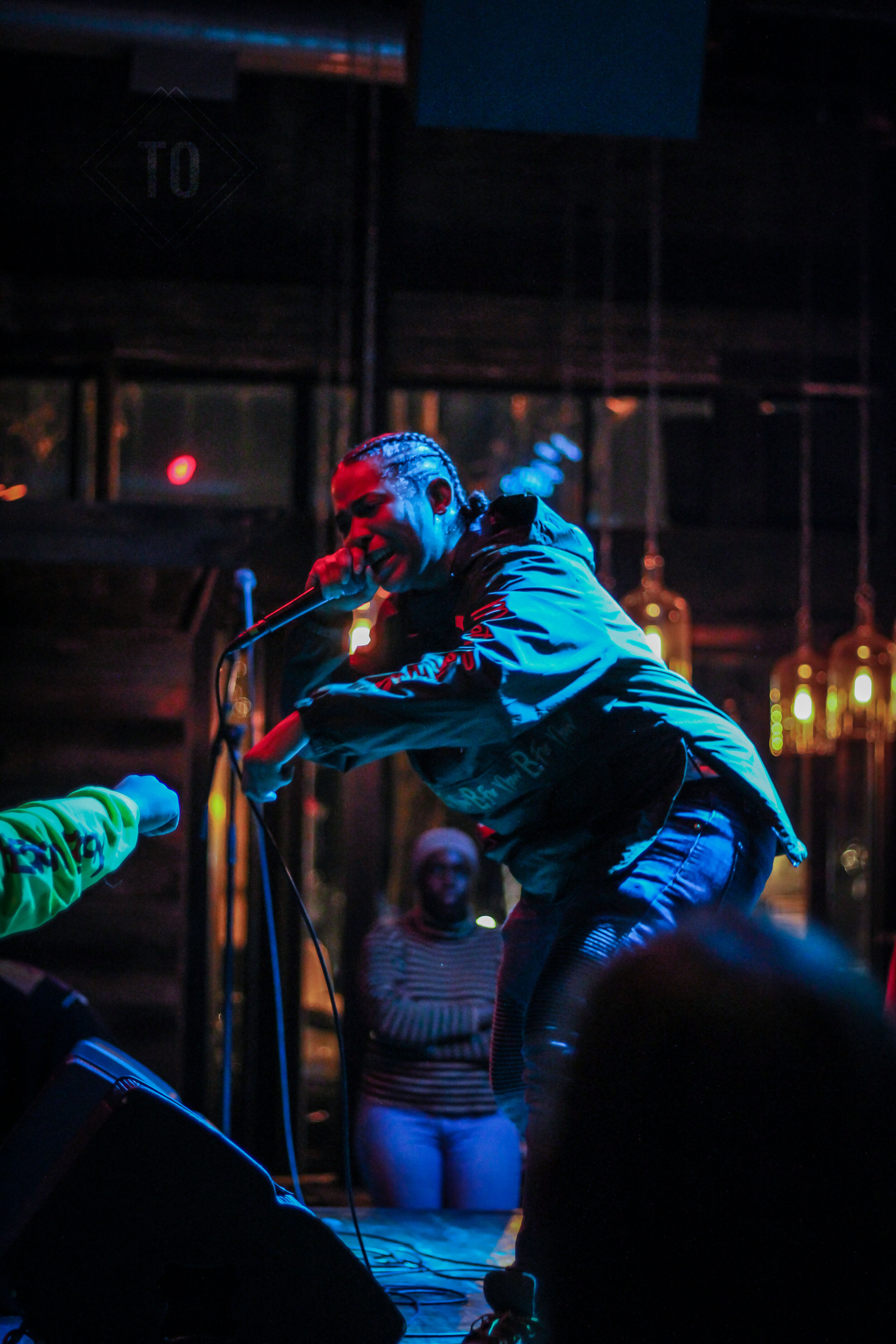 Performer singing into a microphone on stage under colorful lighting at a concert venue, with audience members visible in the background.