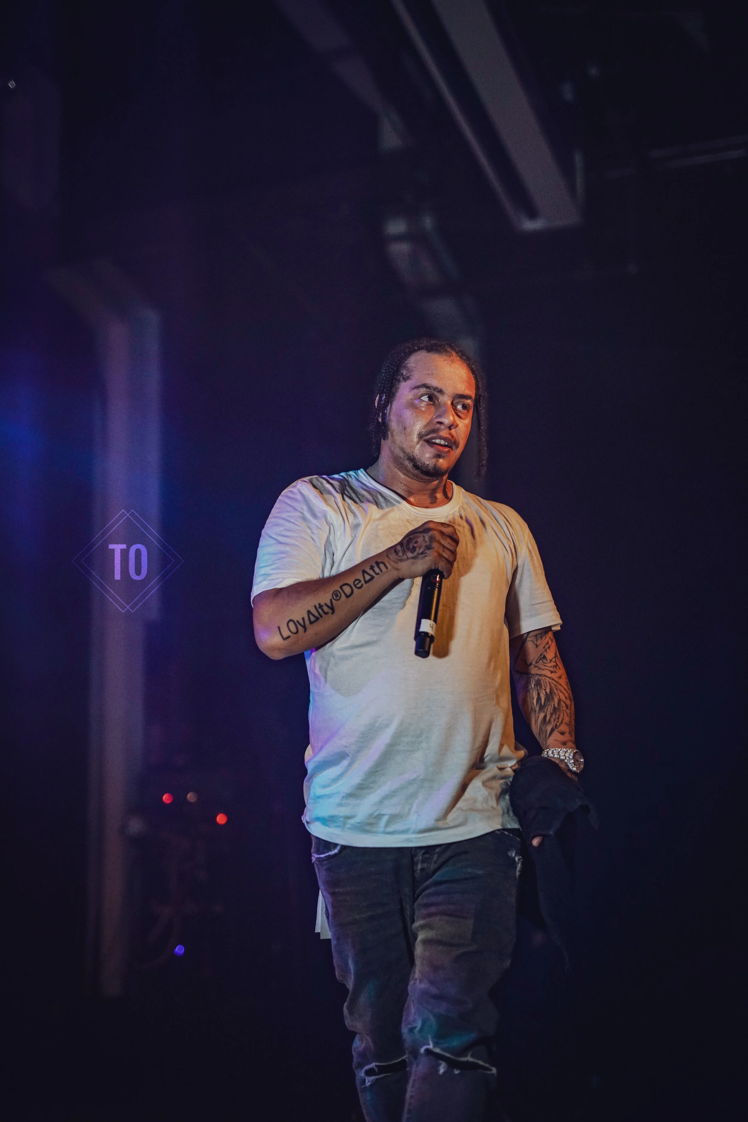 Man with tattooed arms holding microphone, wearing a white t-shirt, on stage during performance