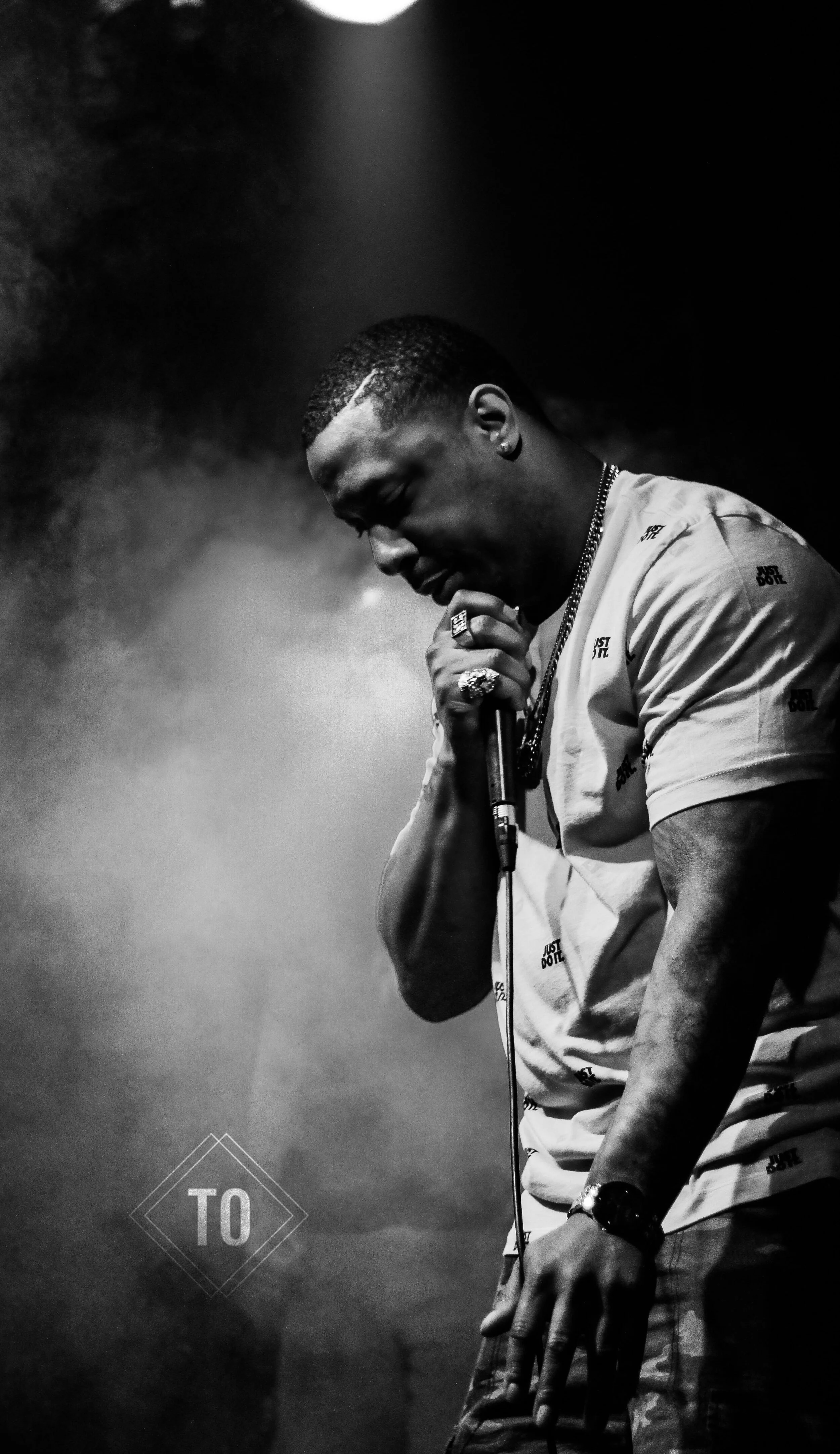 A black and white photo of a male singer performing on stage, holding a microphone with both hands, eyes closed, and head slightly bowed. He is wearing a light-colored t-shirt with small printed text, multiple rings, a watch, and a chain. The stage l