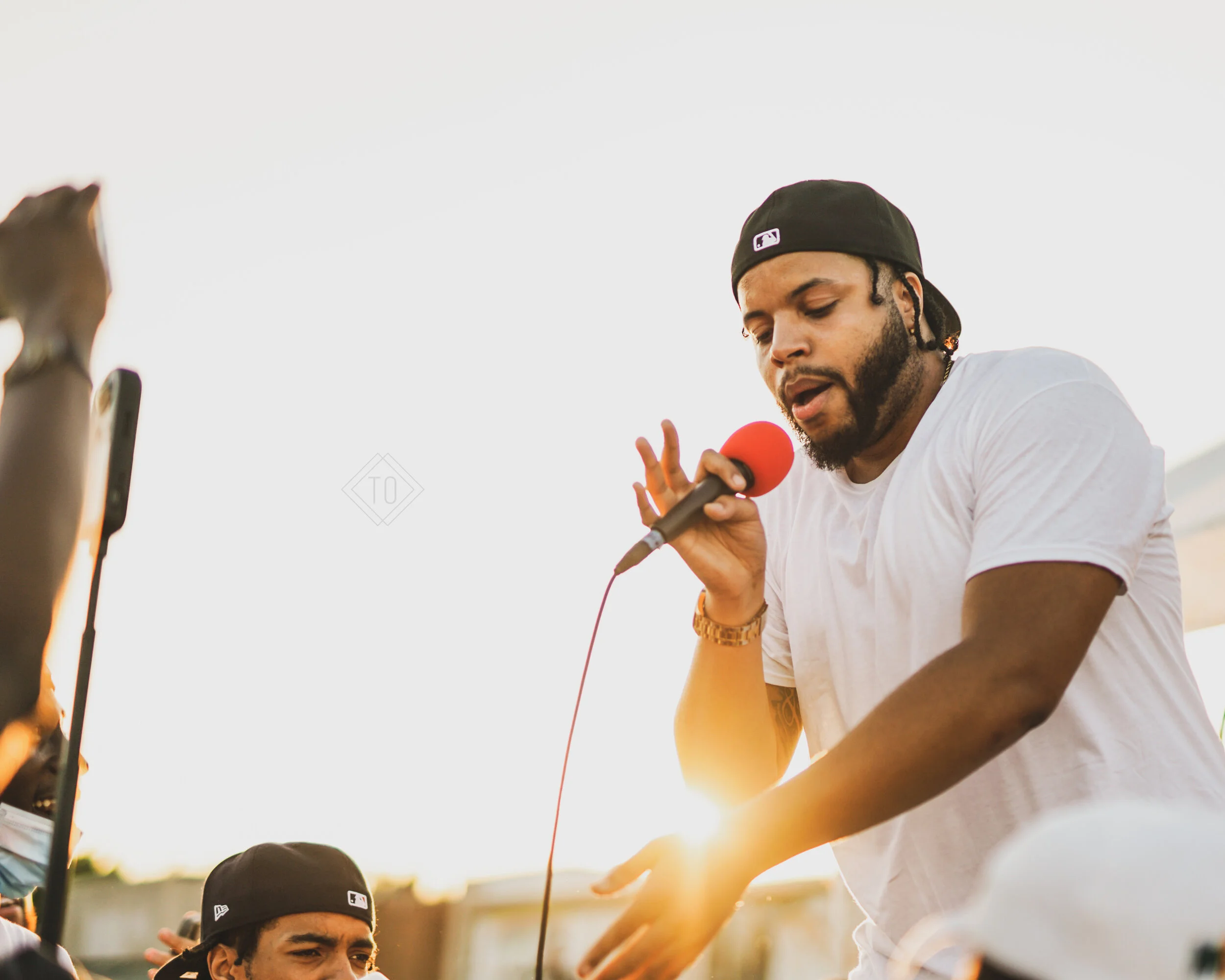 A man in a black baseball cap and white t-shirt holding a microphone with a red windscreen, performing at an outdoor event at sunset.