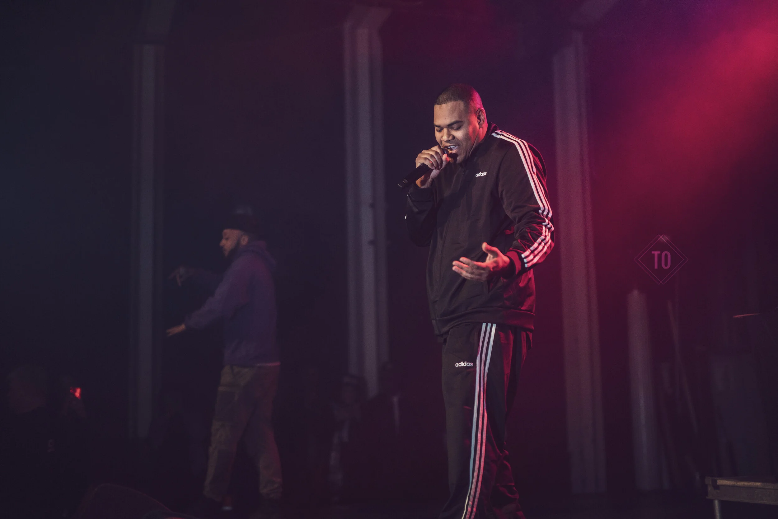 A man in black Adidas tracksuit singing into a microphone on a stage with red lighting.
