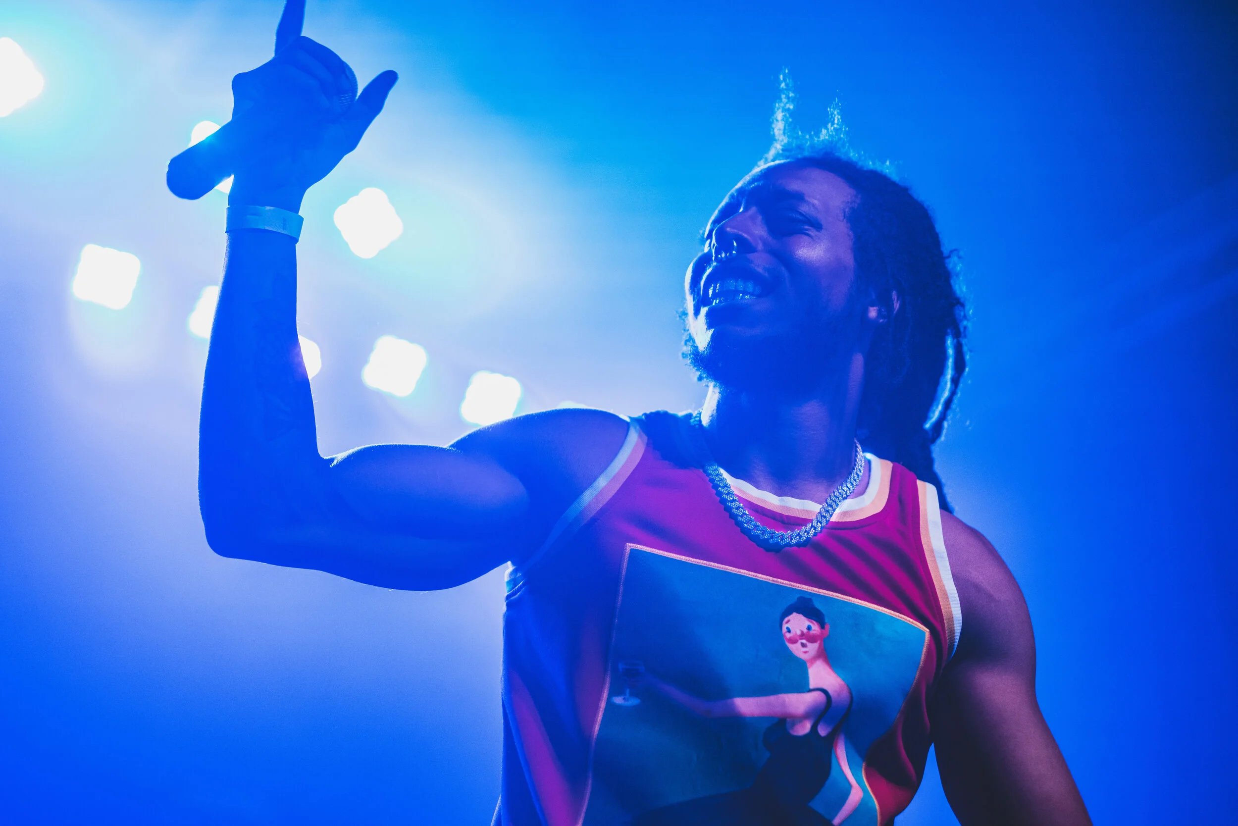 A man with dreadlocks smiling and flexing his bicep on stage under blue lighting, wearing a colorful sleeveless shirt with a graphic design and jewelry.