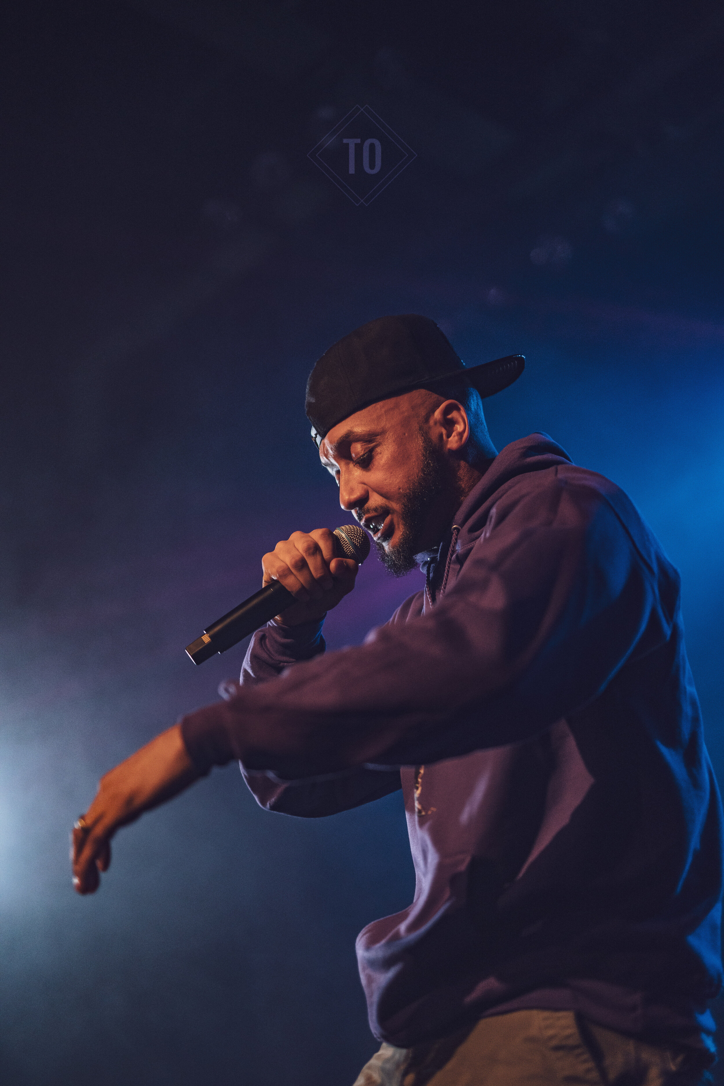 A male performer passionately singing into a microphone on stage, wearing a dark hoodie and a black cap, with colorful stage lights creating a dramatic atmosphere.