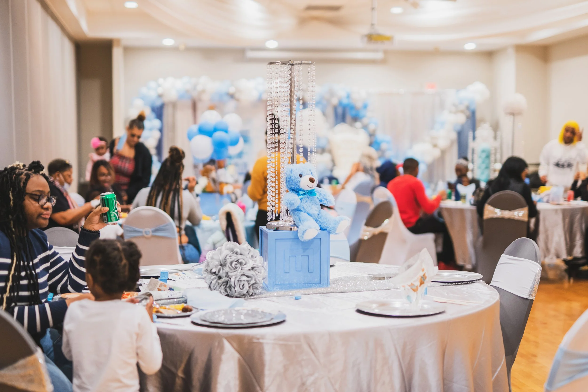 A children's birthday party celebration with a blue and white teddy bear centerpiece, surrounded by balloons and children and adults seated at decorated tables.