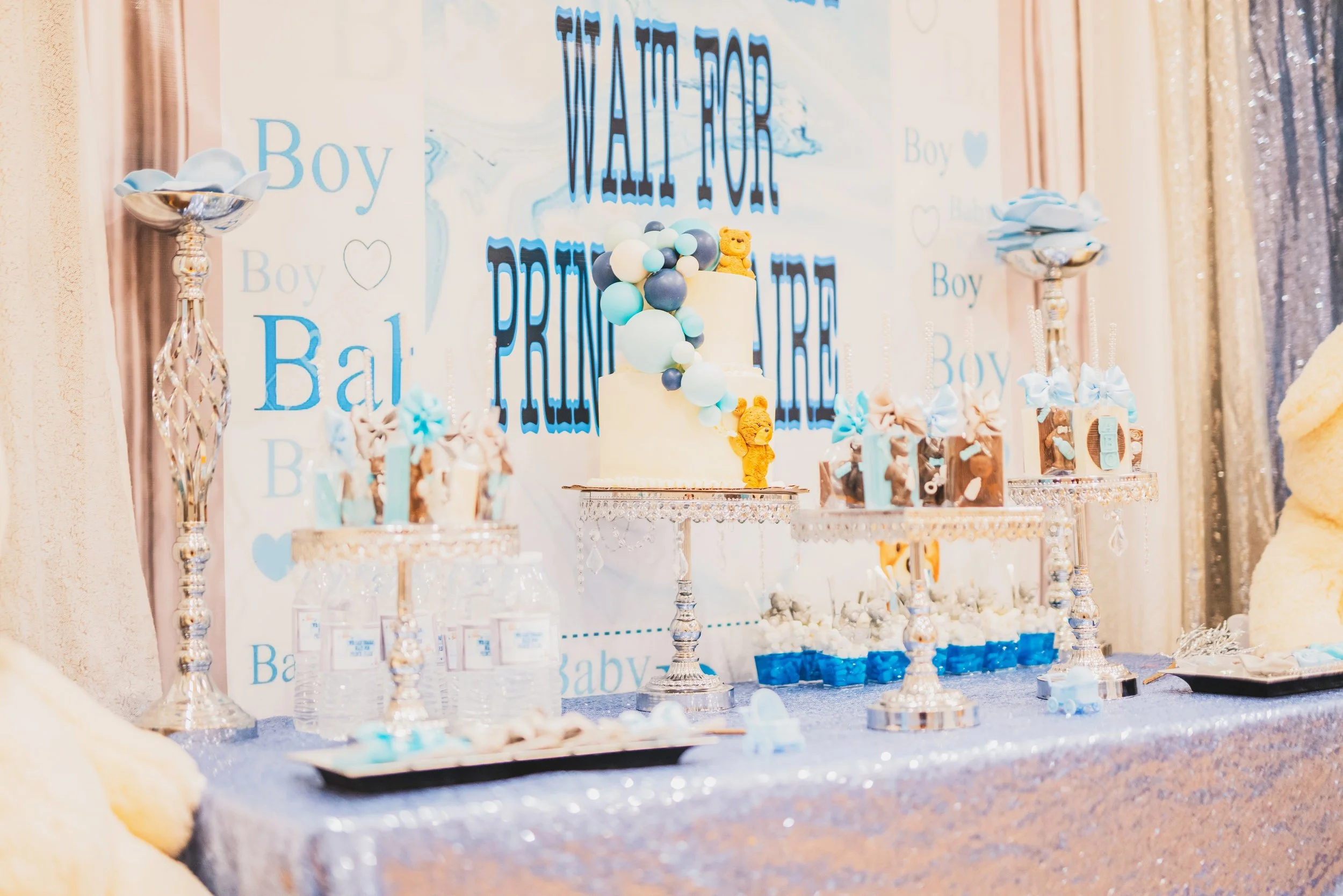 Baby shower dessert table with blue and white decorations, a two-tier cake decorated with blue balloons and teddy bears, balloons, and themed cupcakes, with a backdrop that says 'WAIT FOR PRINCE' in blue lettering.