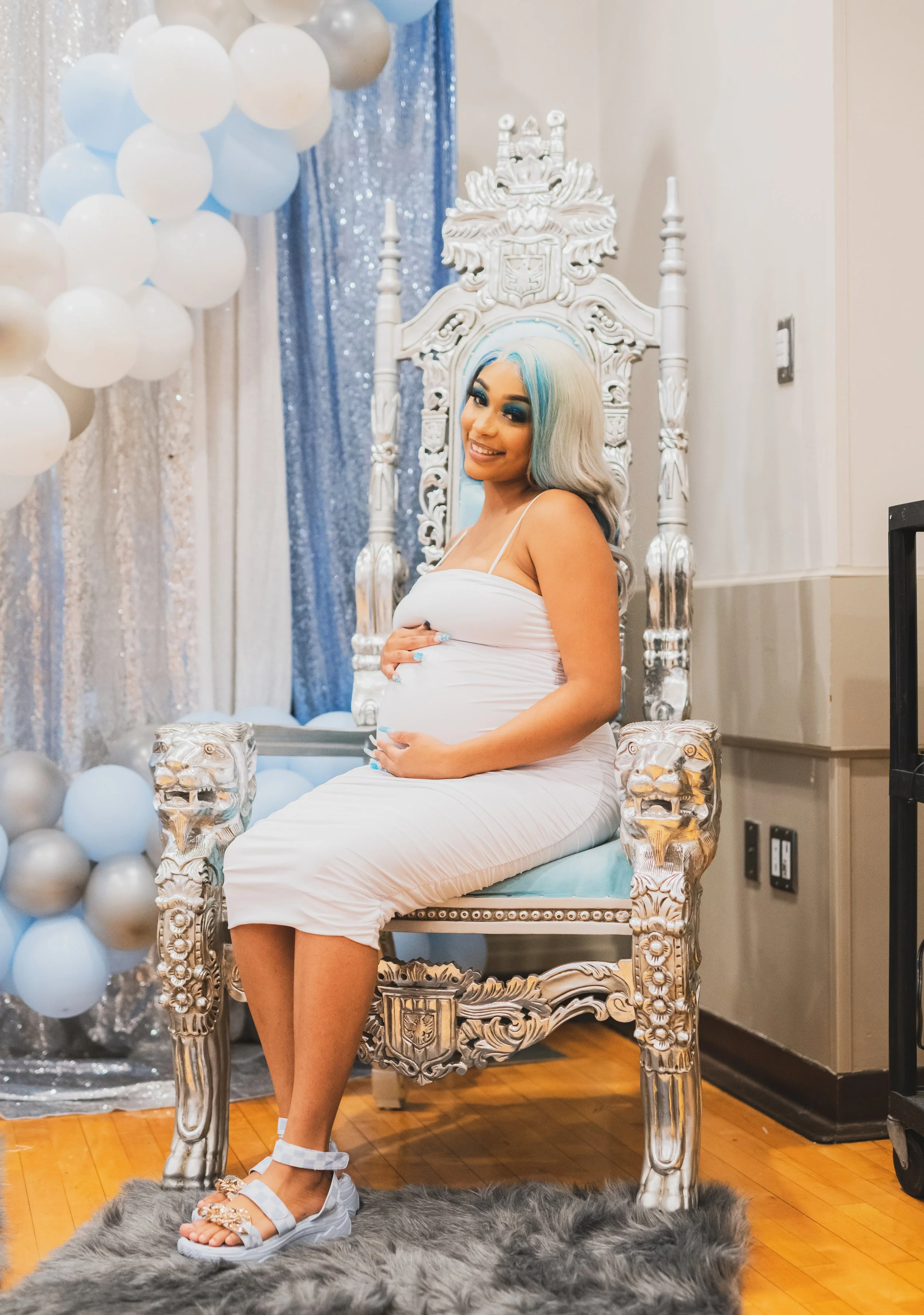 A pregnant woman sitting on a silver throne with intricate carvings, smiling and holding her baby bump, in front of a backdrop of balloons and shimmering curtains.