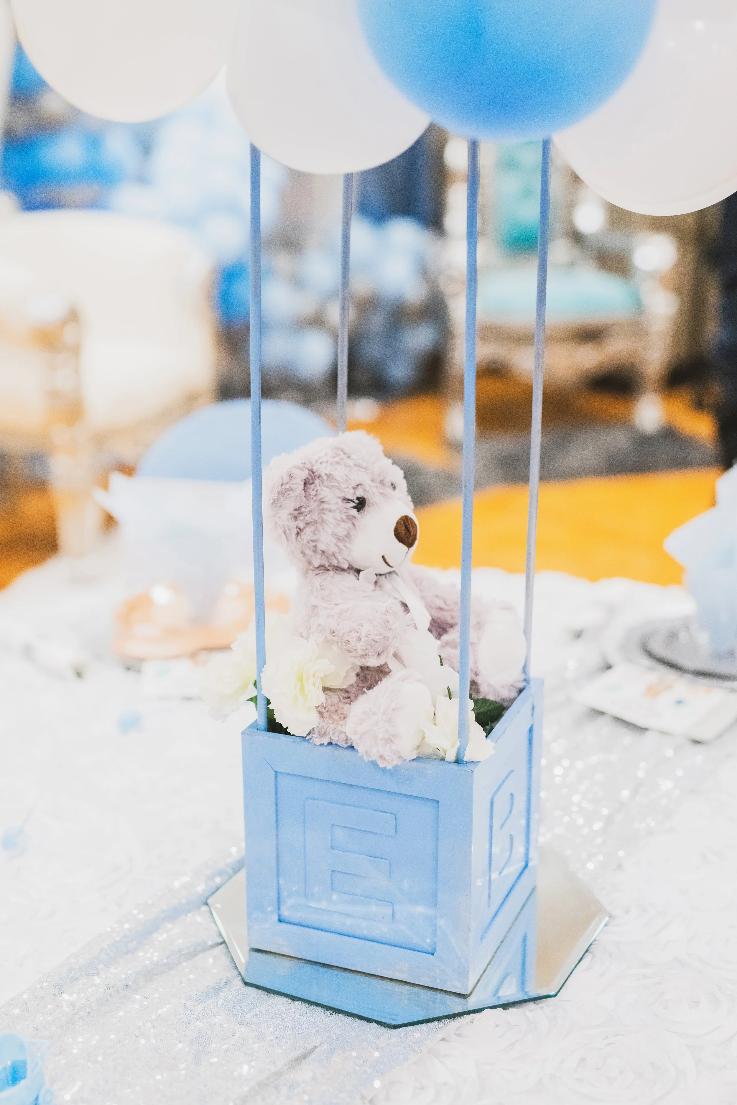 A plush teddy bear sitting inside a blue hot air balloon basket decoration on a table at a party or baby shower, with balloons overhead and other blurred decorations in the background.
