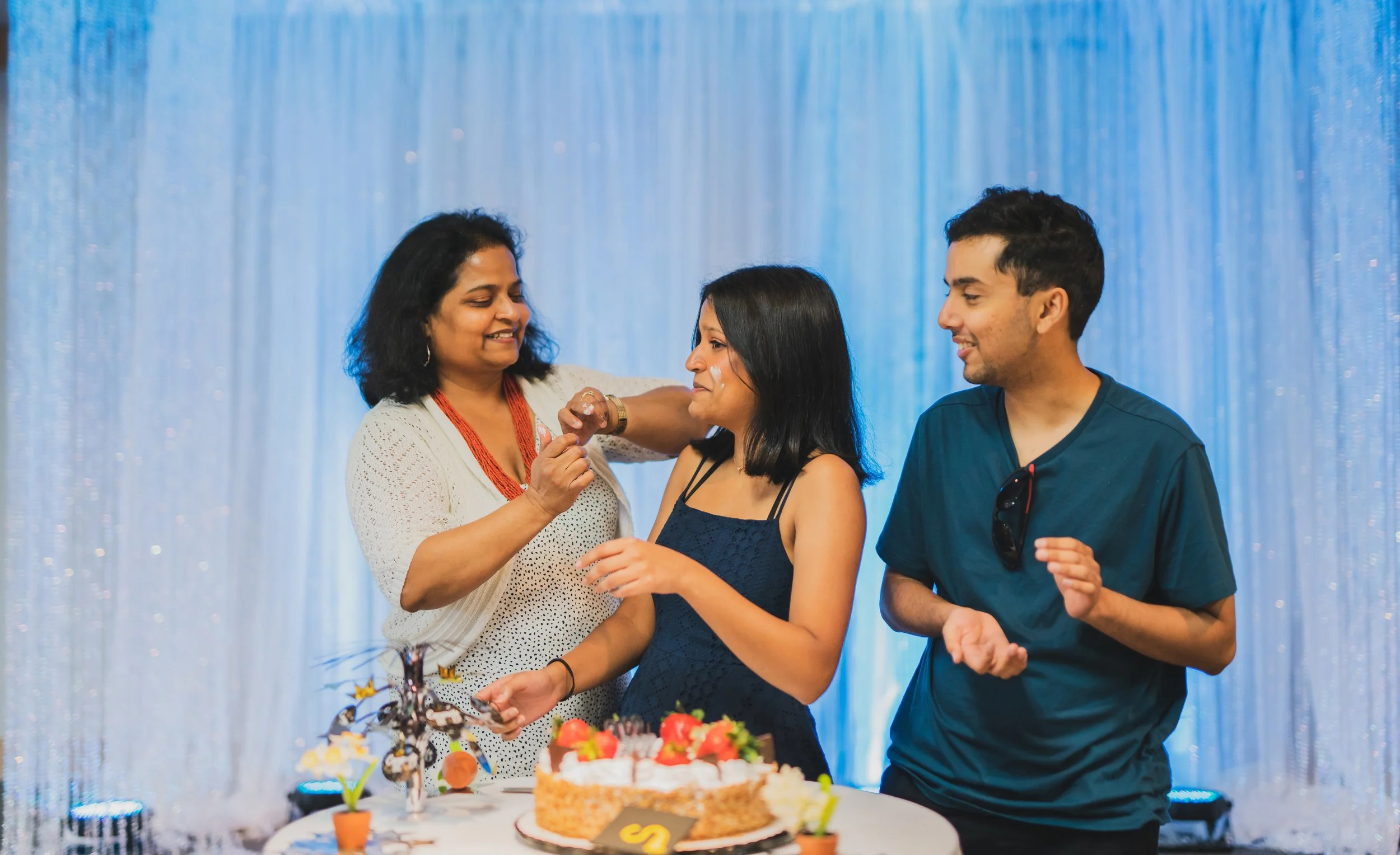 A birthday celebration with three people around a table with a cake, balloons, and decorations. A woman is receiving a gift or a card from another woman, while a young man looks on happily. The background features shiny blue curtains.