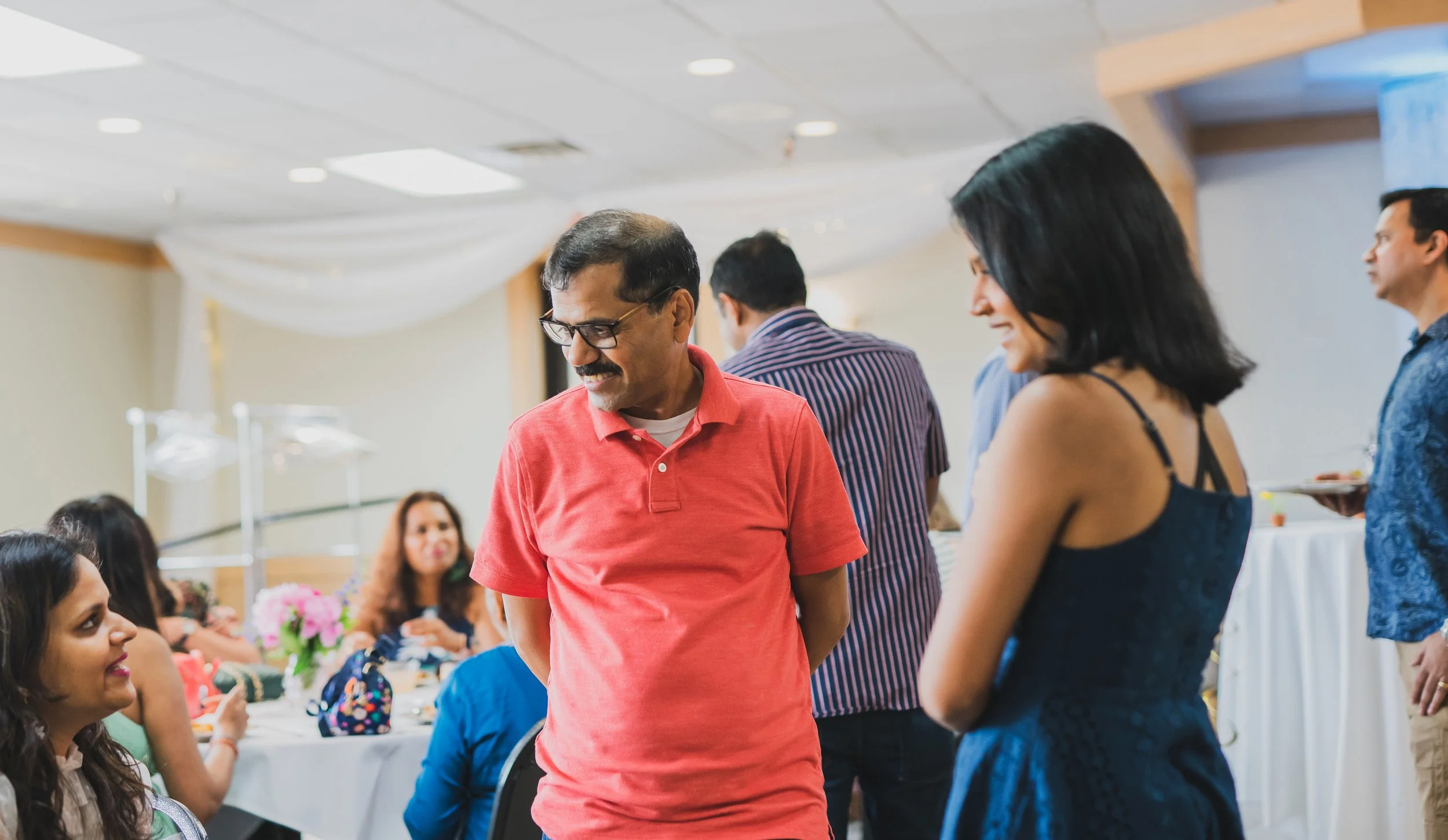 People socializing at a gathering in a banquet hall, with decorations, tables, and floral arrangements visible.