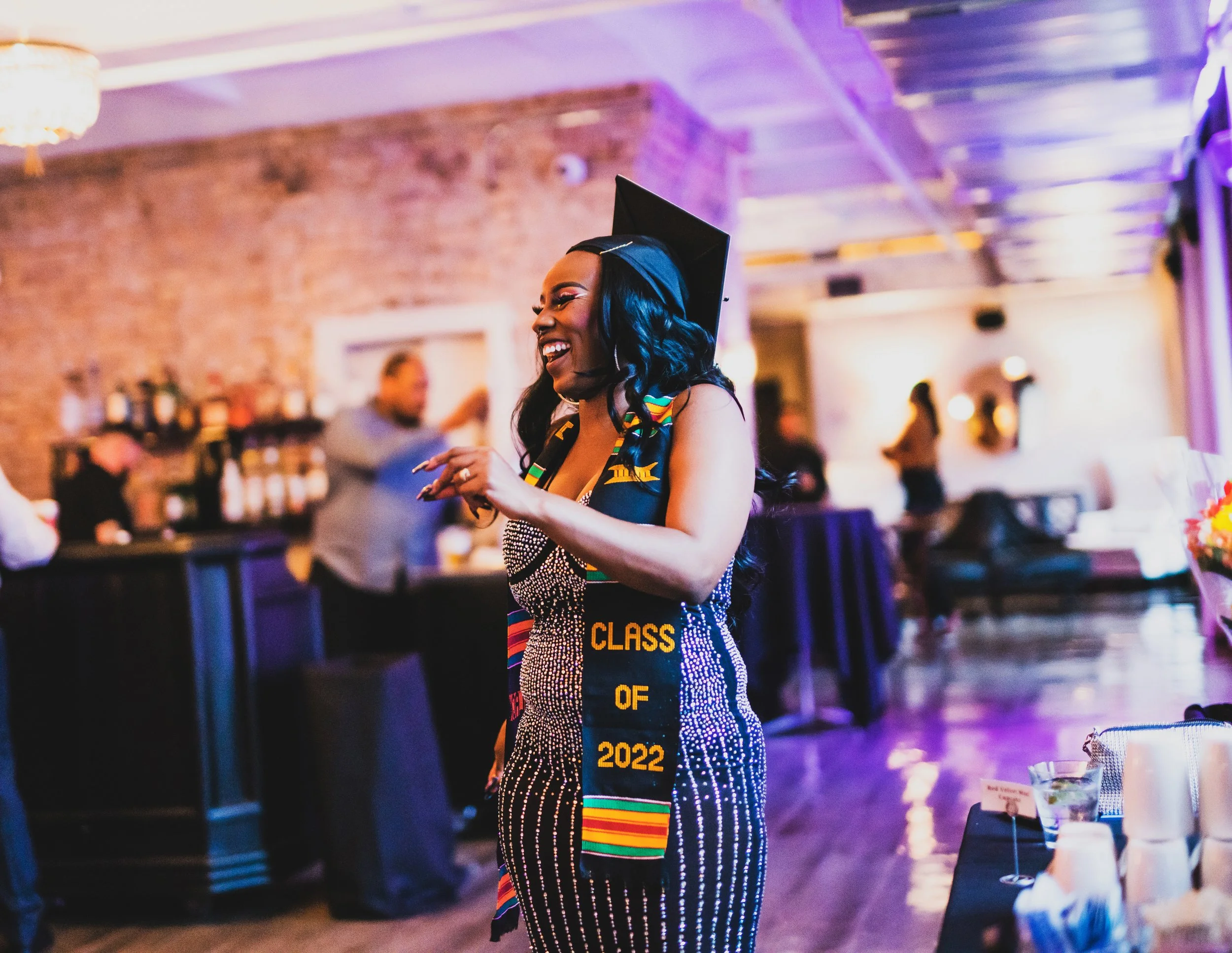 A woman at a graduation celebration, wearing a black mortarboard and a colorful stole that reads 'Class of 2022'. She is smiling and appears to be happy in a decorated indoor space with guests and a bar in the background.