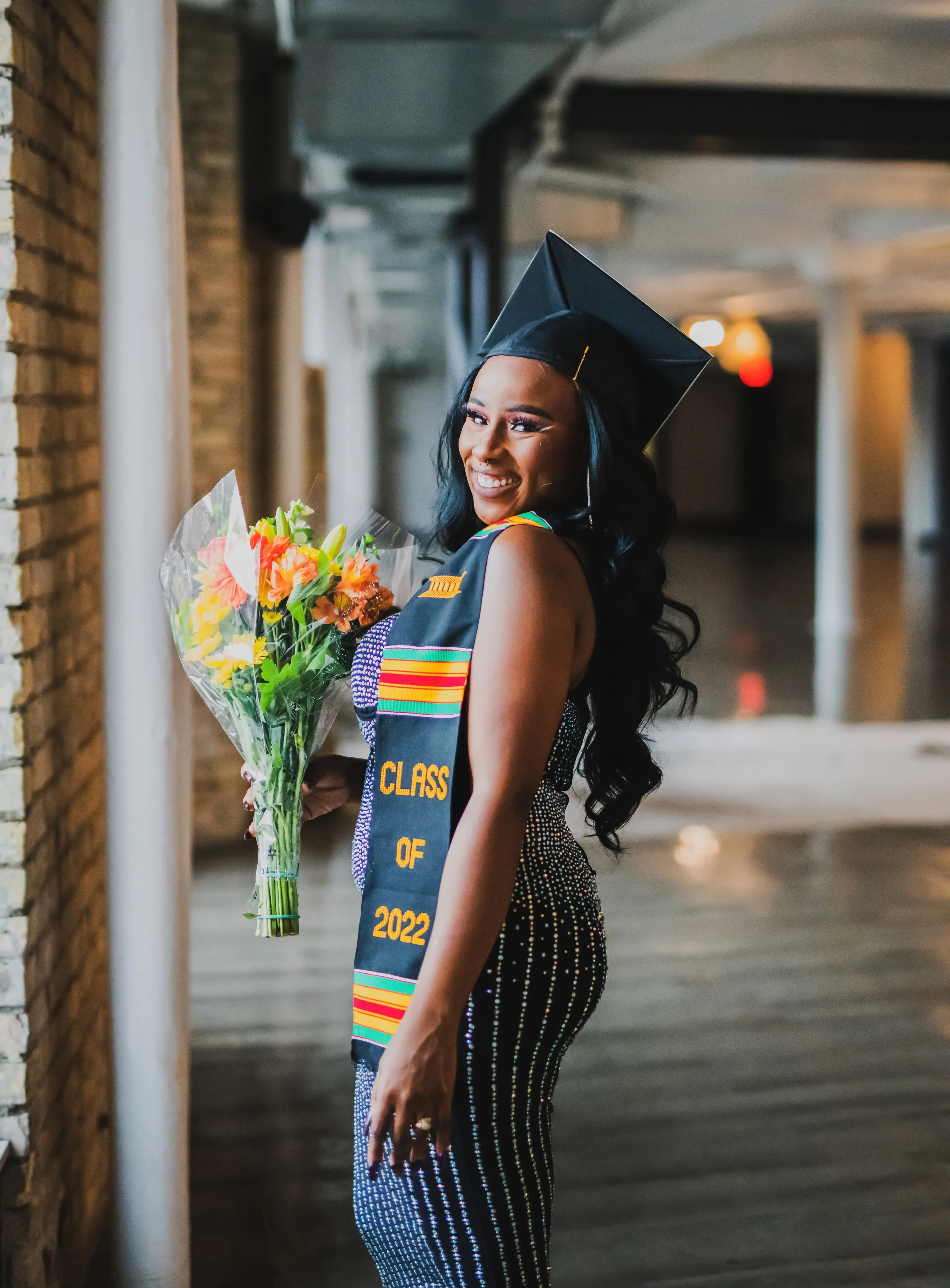 A young woman with long dark hair, dressed in a sparkly gown, holding a bouquet of flowers, wearing a graduation cap and stole that reads "Class of 2022," smiling and standing in a hallway.