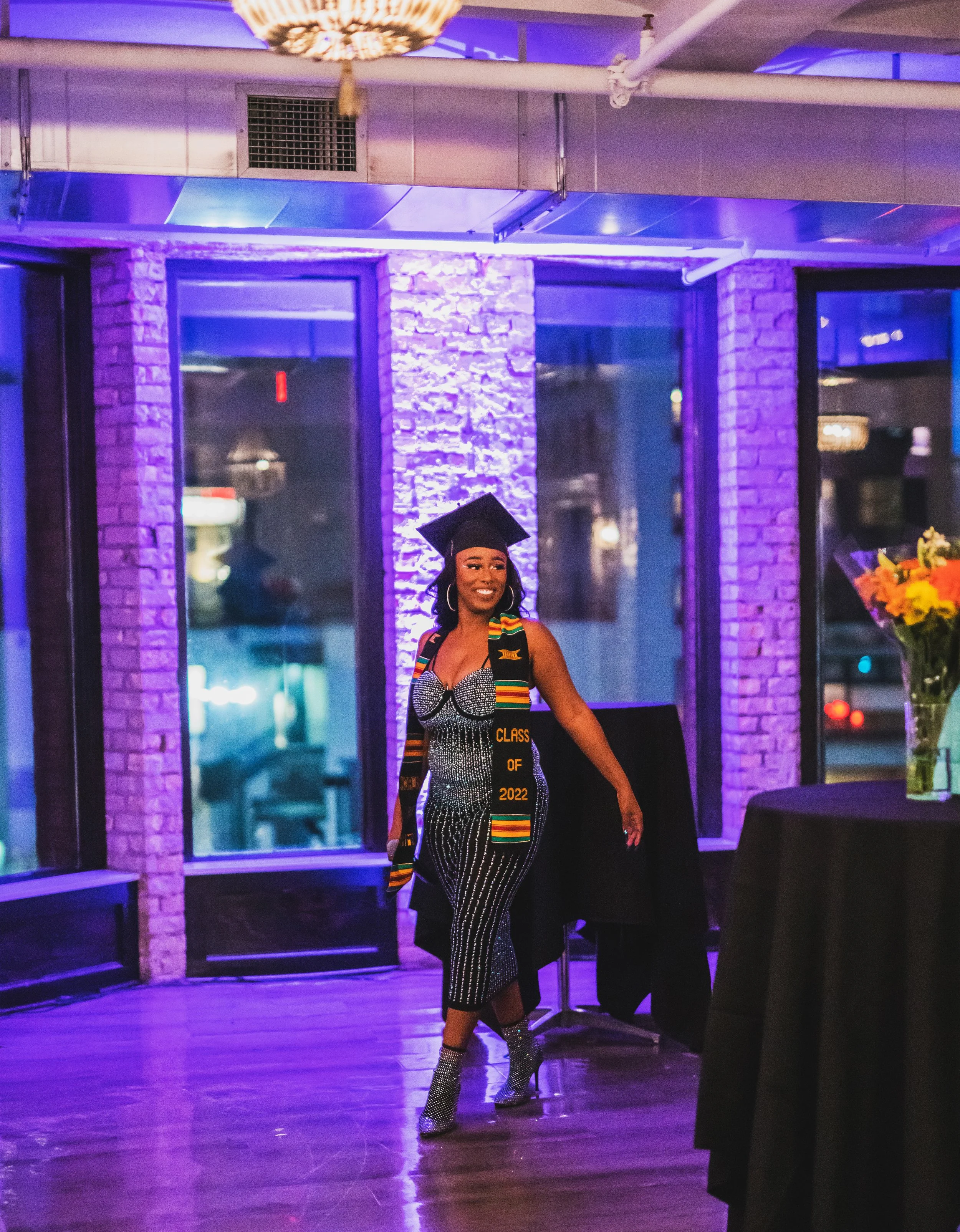 Graduate woman in a black graduation cap and gown, wearing a kente stole and a dress, smiling at a celebration in a decorated venue with purple lighting.