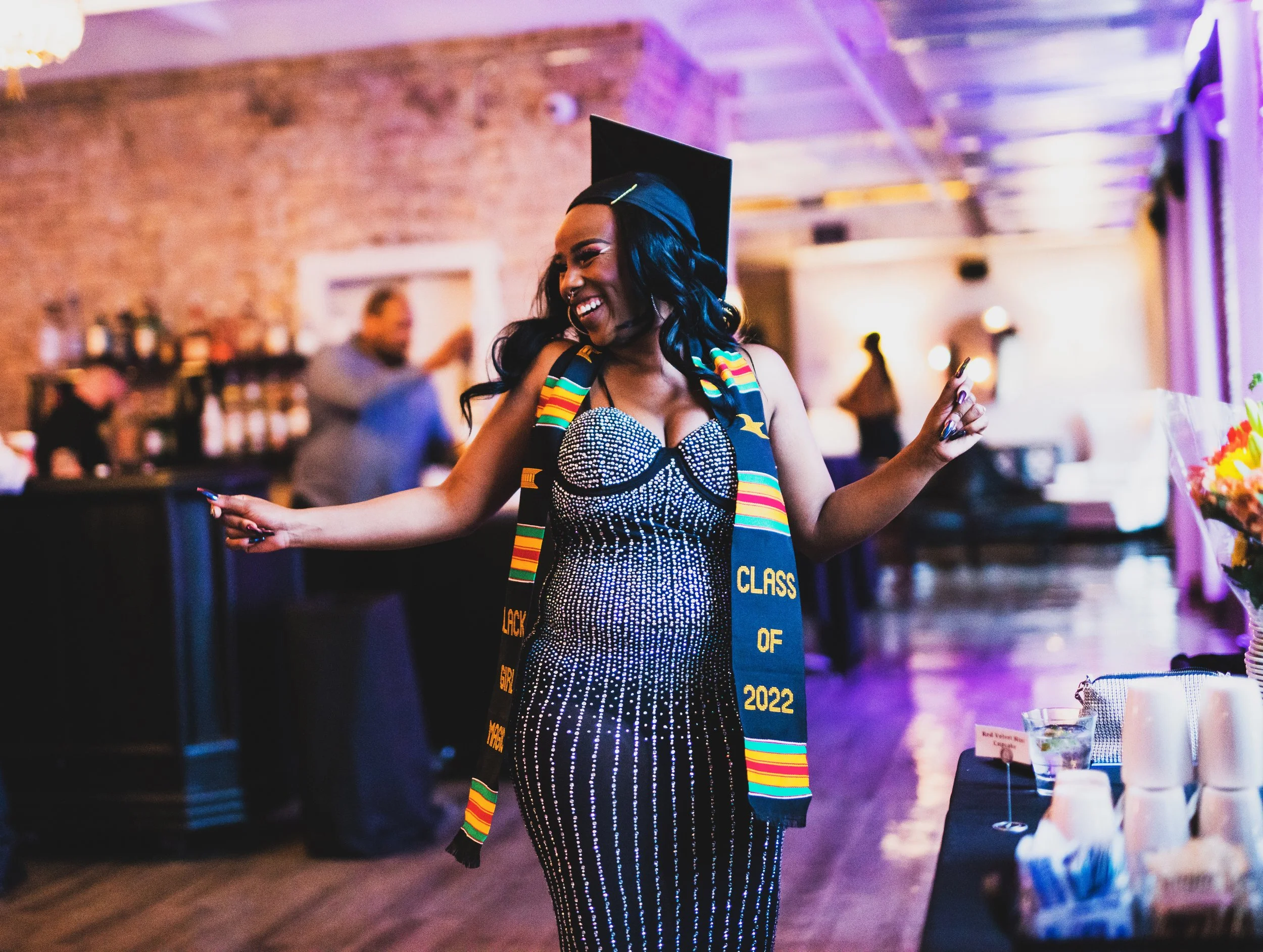 A woman in a black and white beaded dress celebrating her graduation, wearing a cap and a graduation stole that reads 'Class of 2022'. She is smiling and dancing at a celebration event in a dimly lit venue with exposed brick walls.