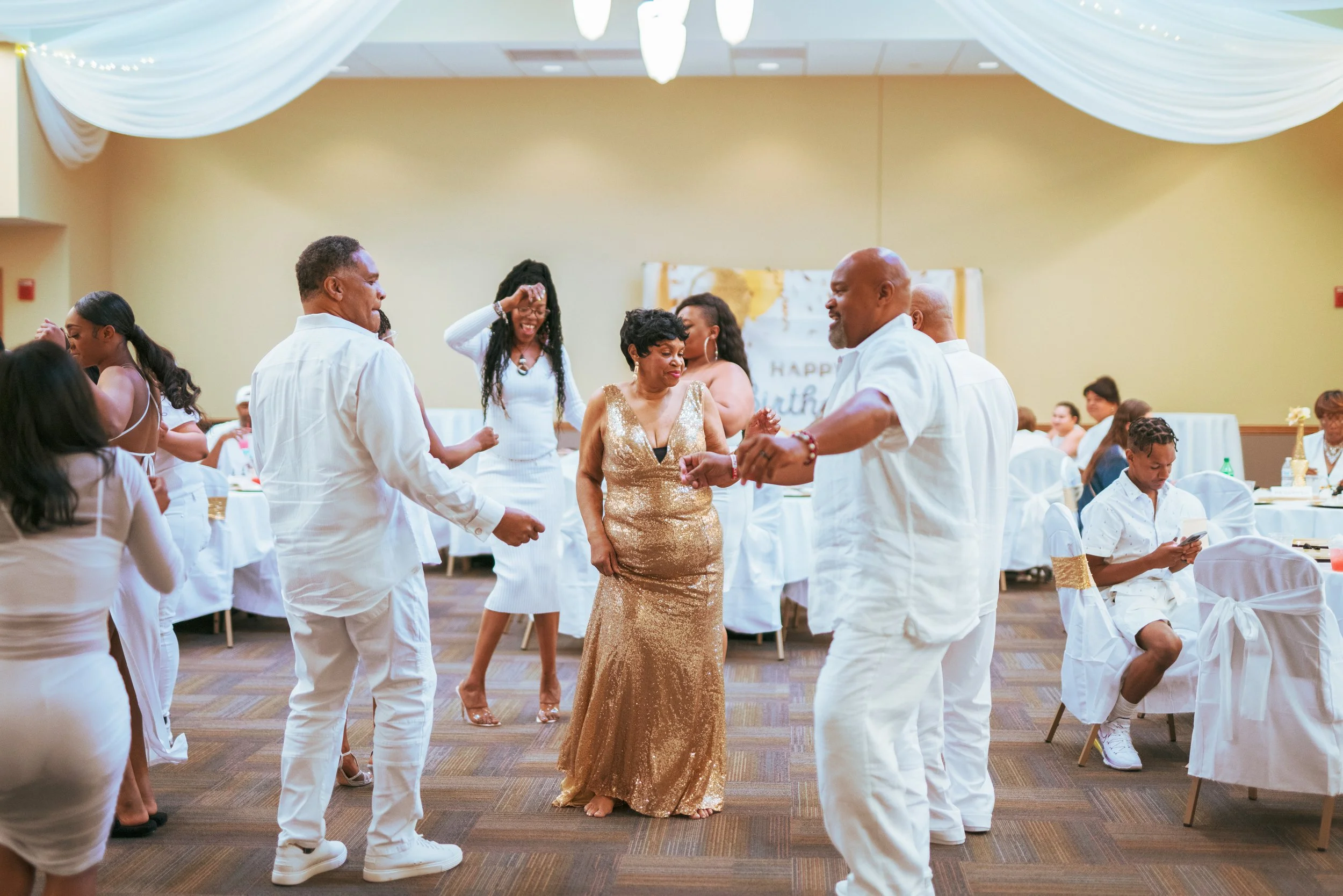 People dancing and celebrating at a party, with some seated and others dancing in a decorated event hall.