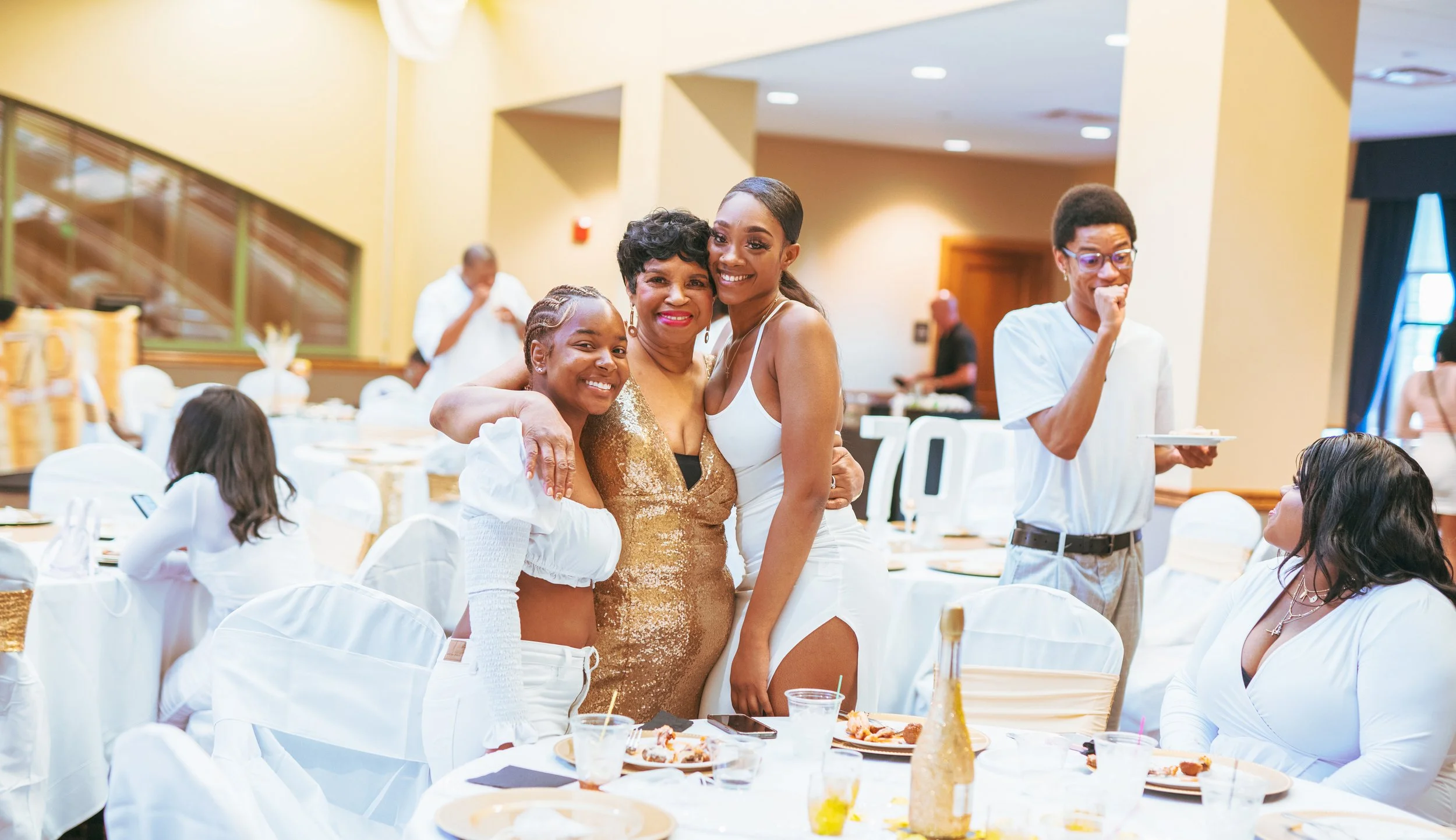 Celebration or party scene with three women hugging and smiling at a banquet table, others seated or standing in the background, in a decorated venue with food and drinks.