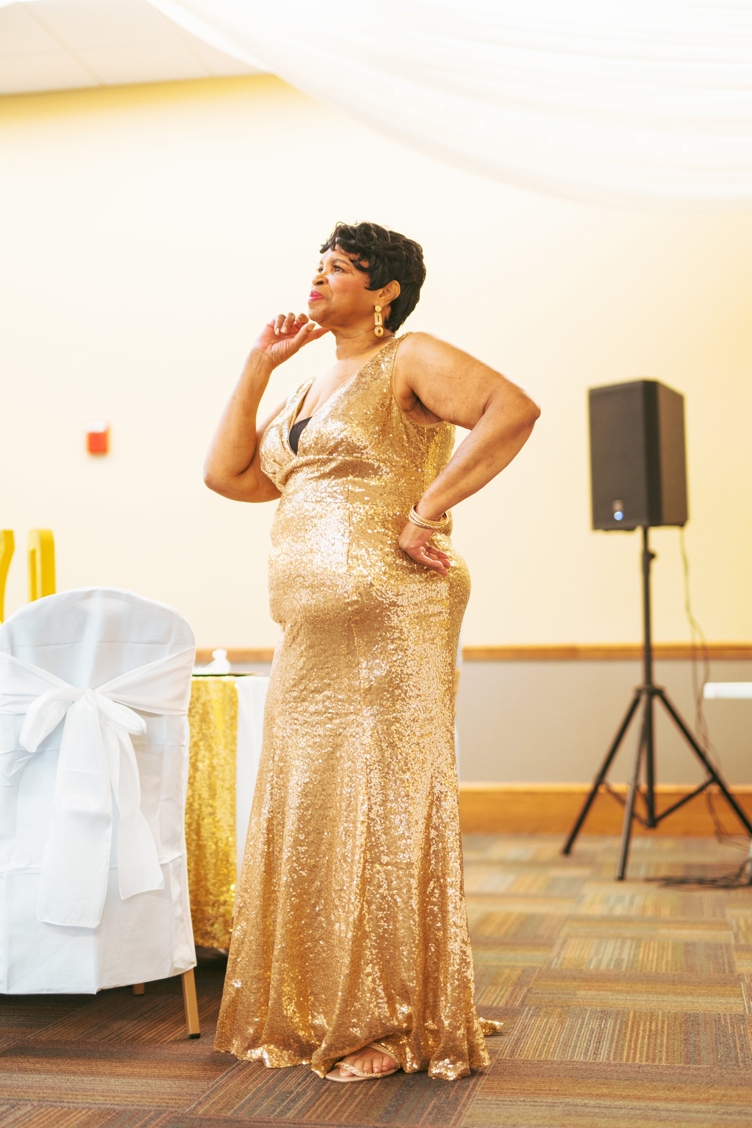 A woman wearing a gold sequin evening gown and gold jewelry standing in a decorated event room.