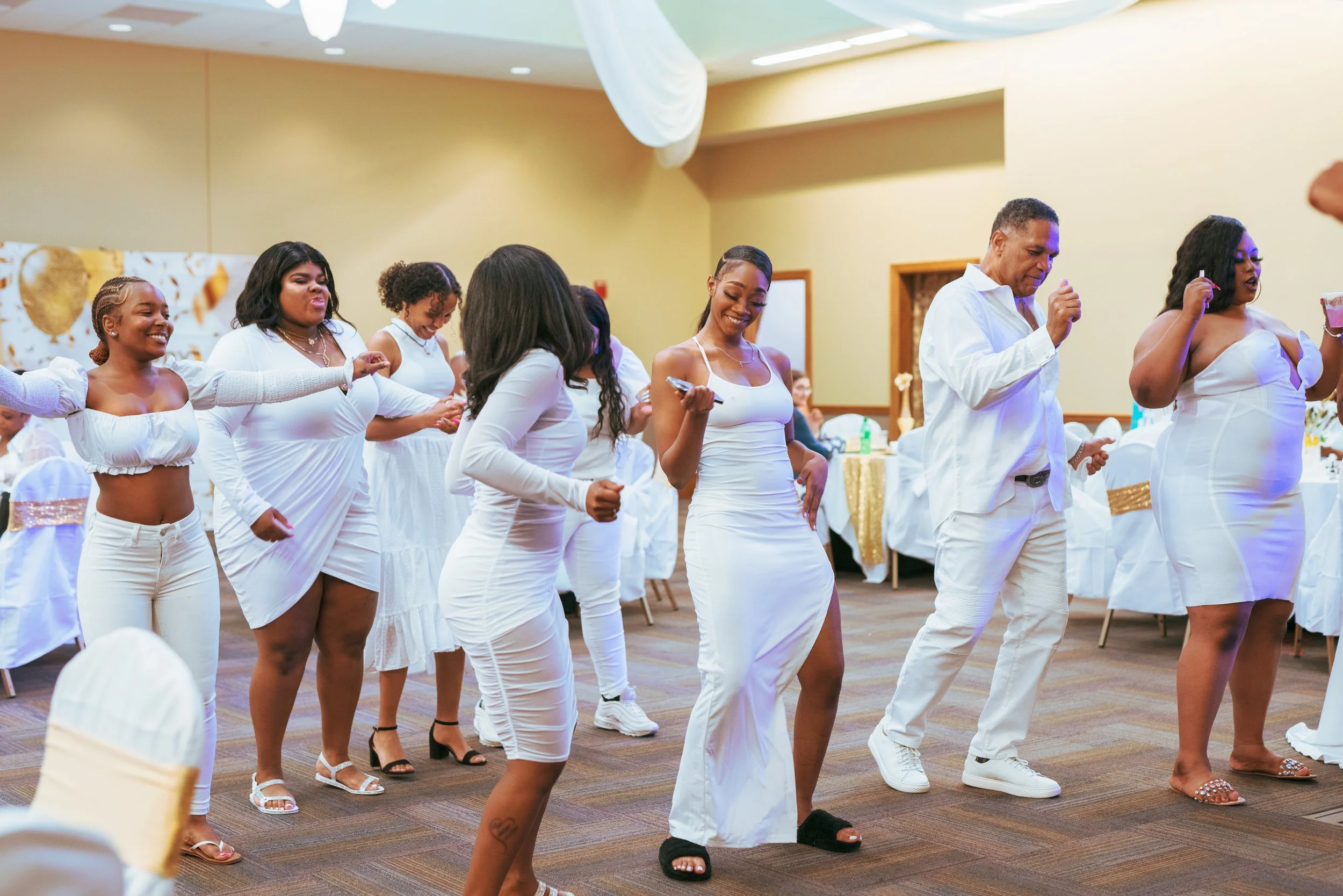 Group of people dancing at a celebration or party, all dressed in white, in a decorated indoor venue.
