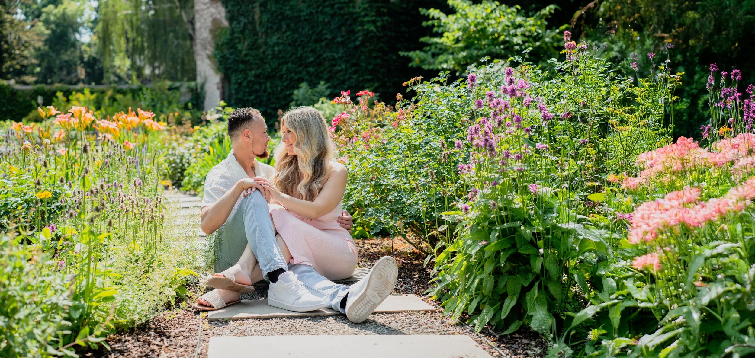 A couple sitting on a garden path surrounded by colorful blooming flowers, gazing into each other's eyes.