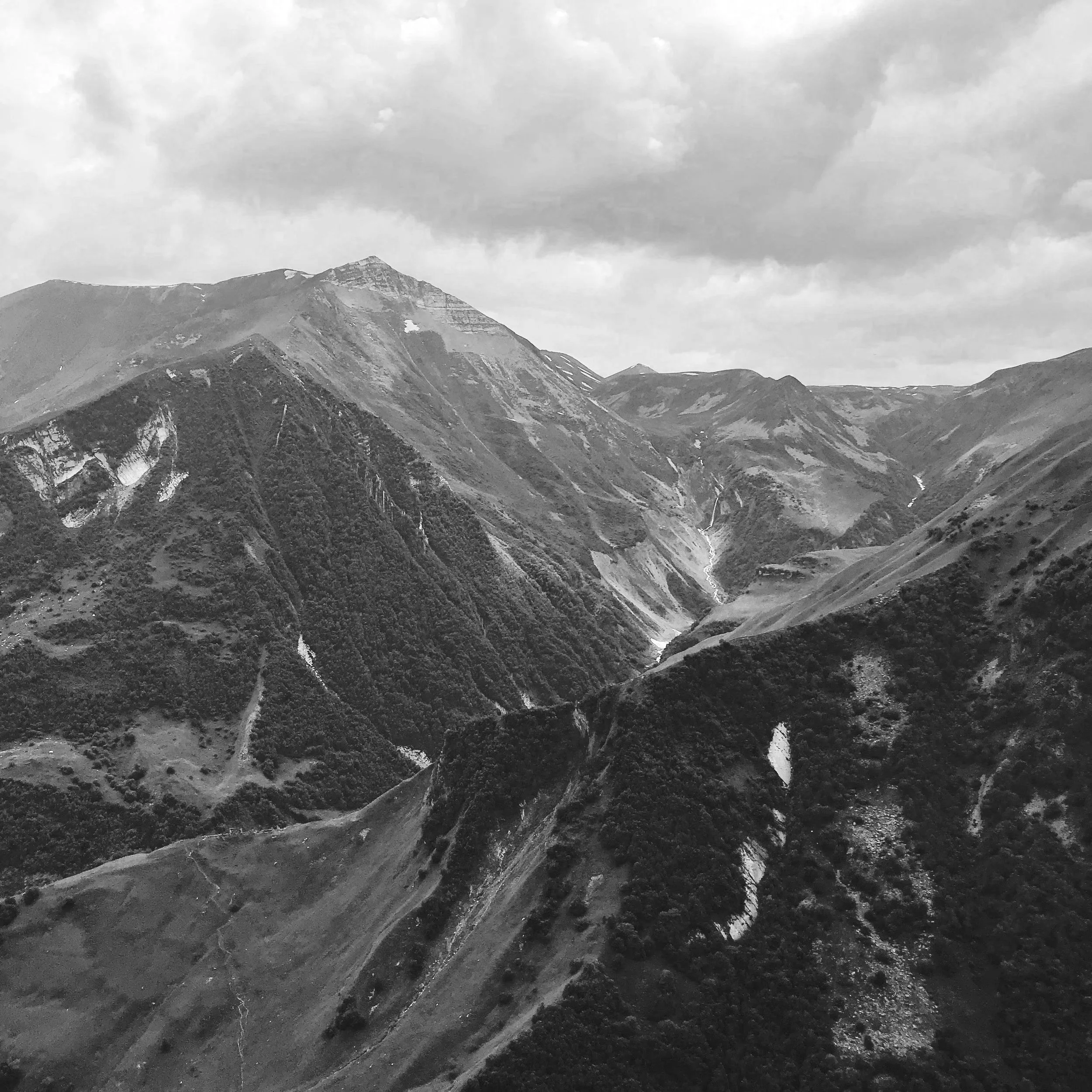 Black and white photo of the Caucasus mountains landscape with peaks, valleys, and ridges under a cloudy sky.