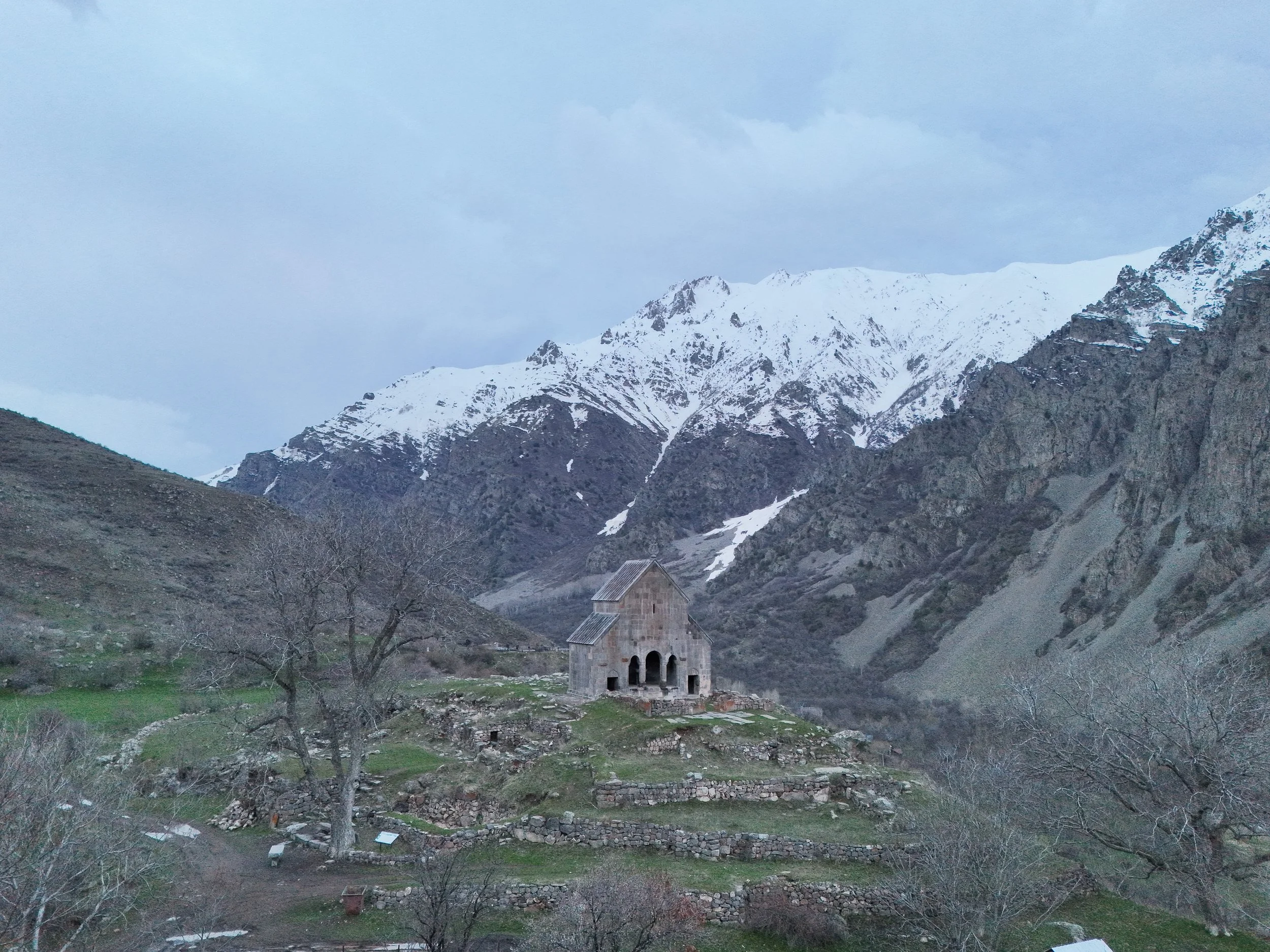 A small medieval stone church with an arched entrance porch sits on a grassy hill among low ruined stone walls. Snow-capped mountains and steep rocky slopes rise behind it under an overcast sky.