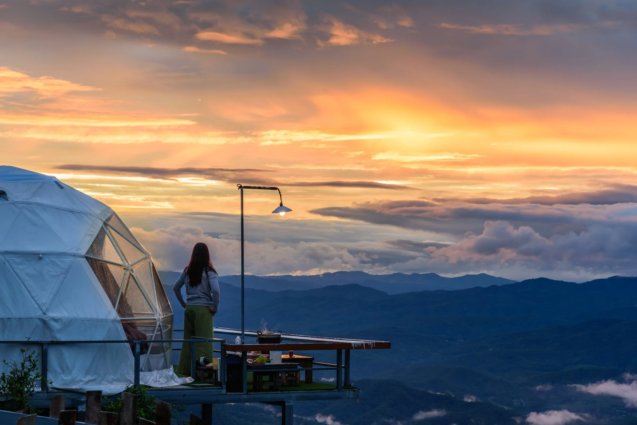 A woman on a balcony overlooking a mountain range during sunset, with a geodesic dome, a streetlamp, and a cooking setup on the balcony showing what our Eco-tourism and wildlife experiences will be like.