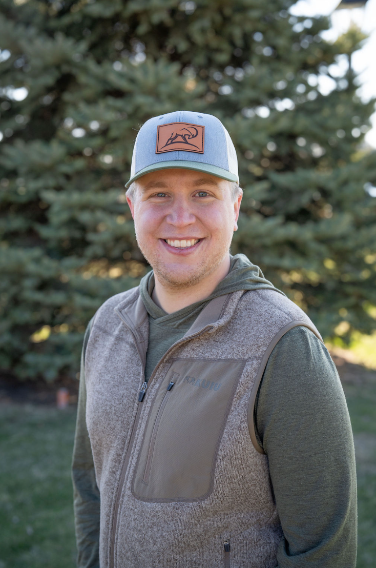 The founder of Wild Giants Conservation Tanner Tripp, wearing a gray and green hoodie, a brown vest, and a blue and gray cap outdoors with green trees in the background.