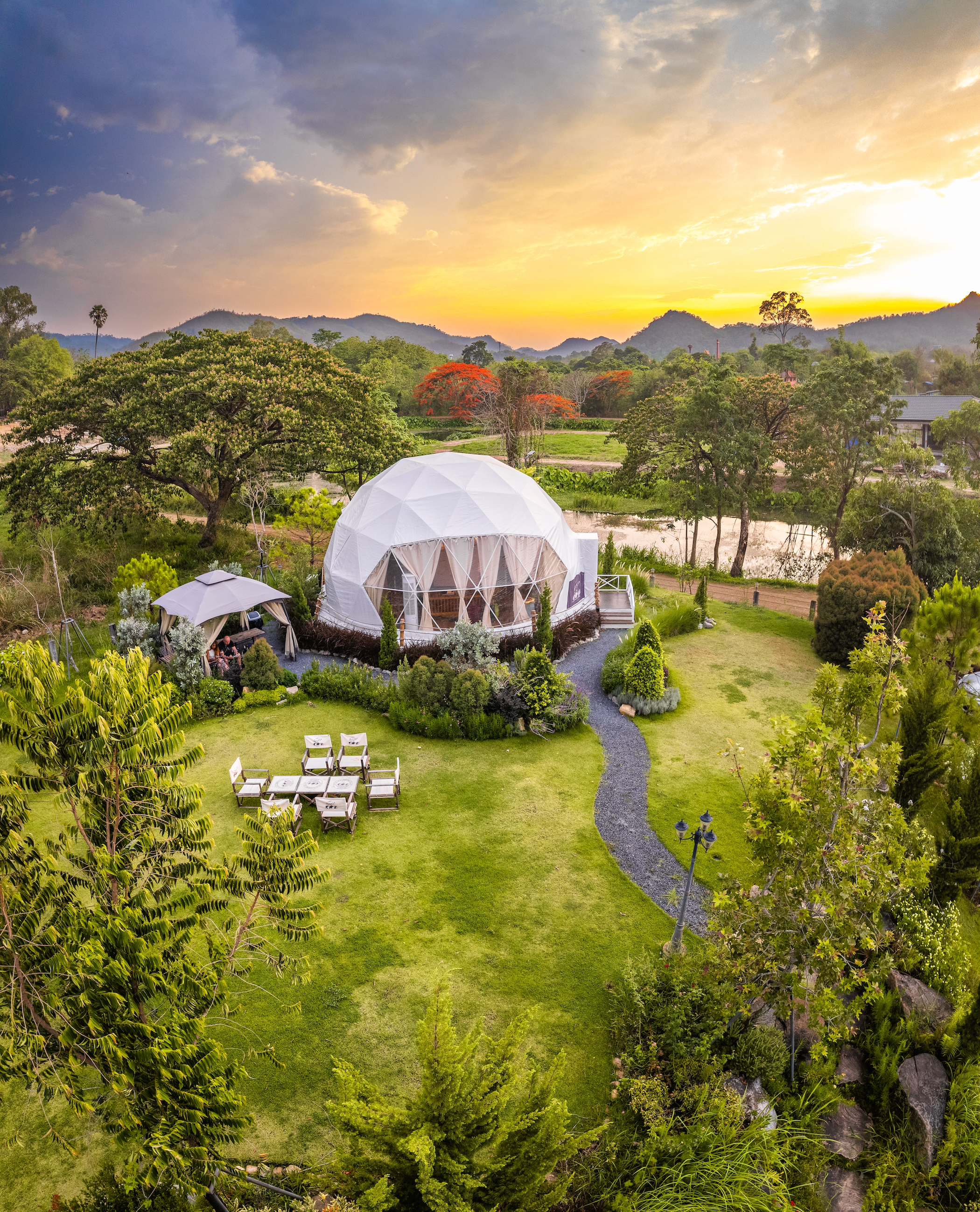 A large geodesic dome in a lush, green landscape during sunset, surrounded by trees, a small pond, a stone pathway, and outdoor seating.