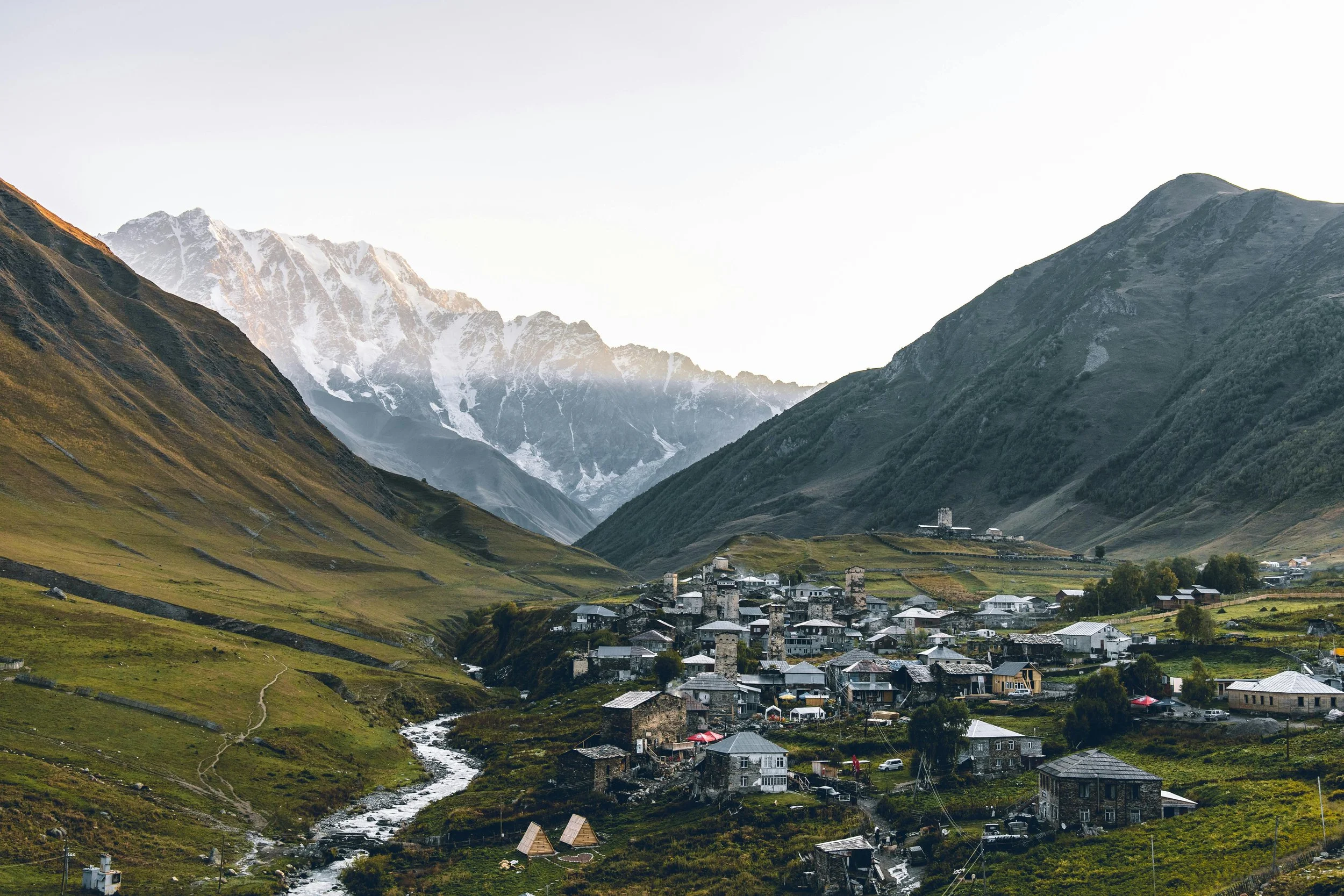 A small village with stone houses and winding roads, situated in a green valley surrounded by tall mountains, some snow-capped, during daylight.