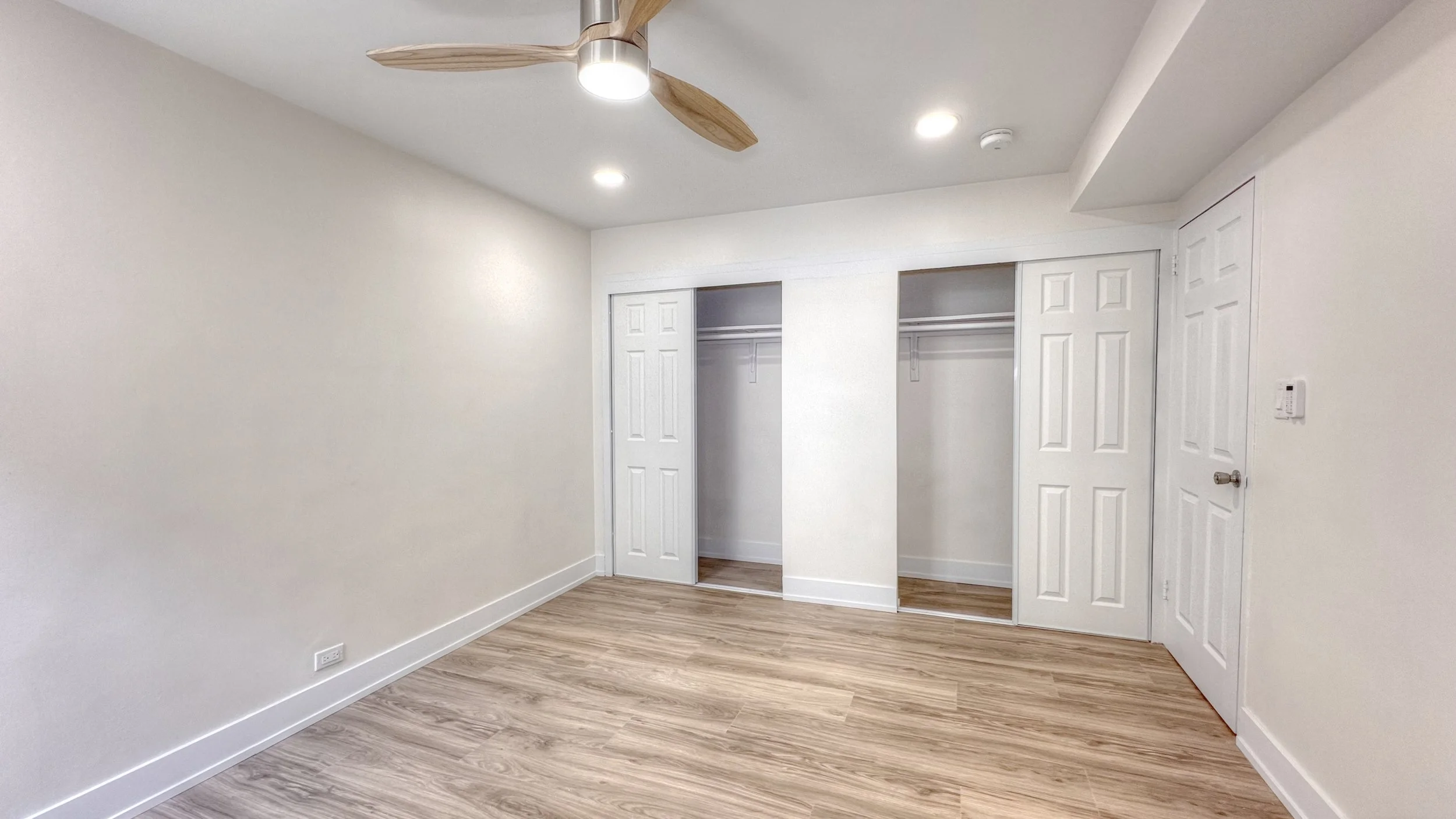 Bright, empty bedroom with light wood-look flooring, white walls, recessed ceiling lights, a modern ceiling fan with wood blades, double sliding-door closets, and a single.jpg