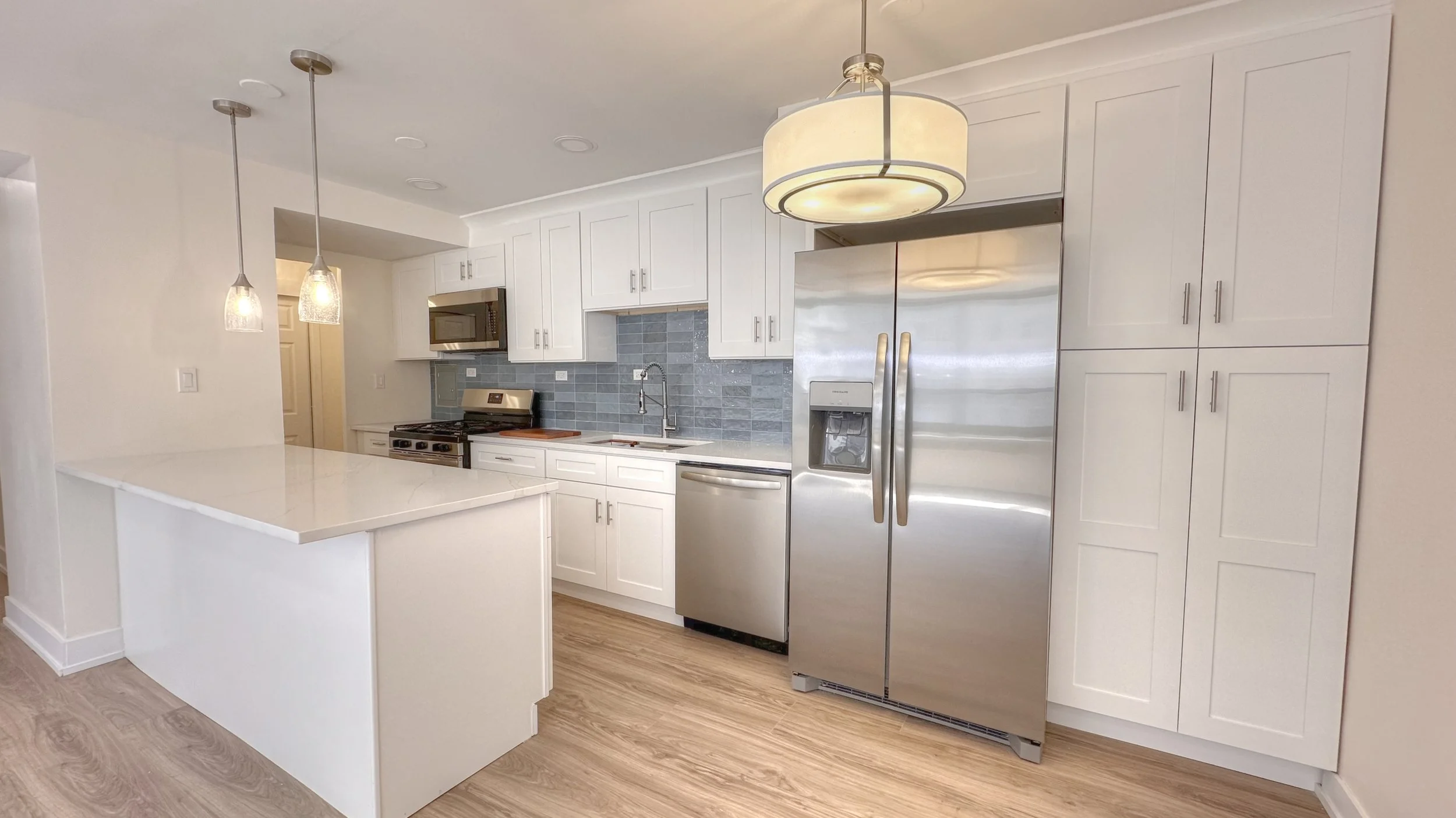 Bright modern kitchen with white cabinets, countertops, stainless steel appliances, blue tile backsplash, breakfast bar seating area, pendant lighting, and wood flooring.jpg
