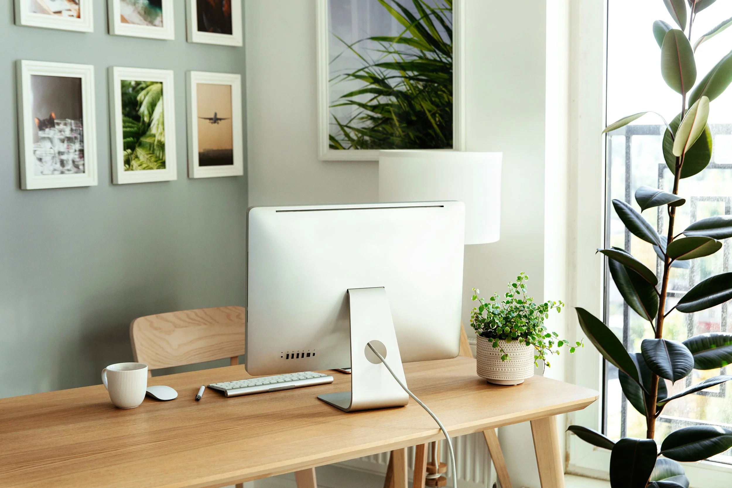 Bright modern home office with a wood desk, desktop computer, framed artwork, and indoor plants beside a large window