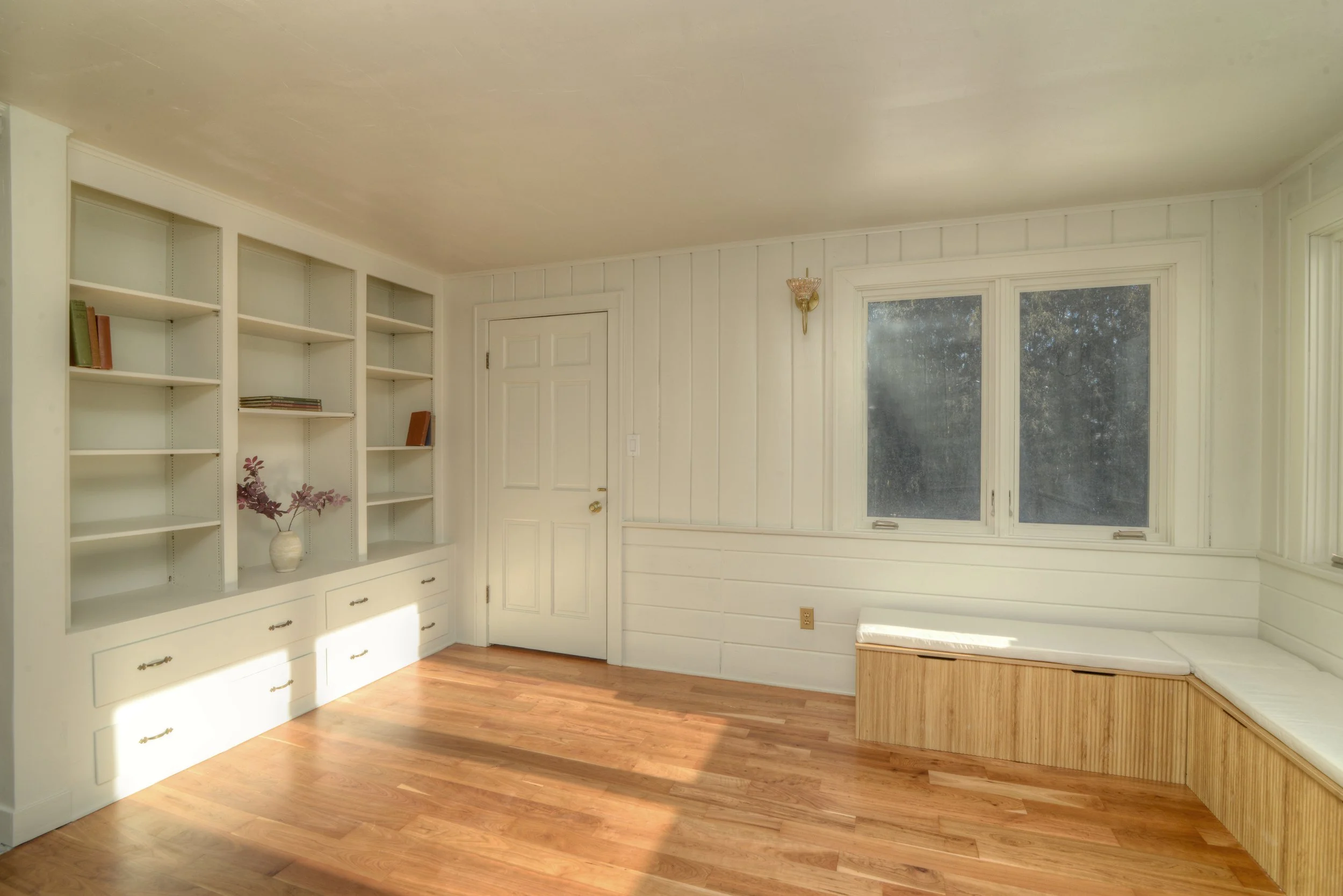 Sunlit room with warm wood floors, white paneled walls, and built-in shelving with lower cabinets. A window seat with storage sits beneath large windows, filling the space with natural light, while a closed interior door anchors the wall.